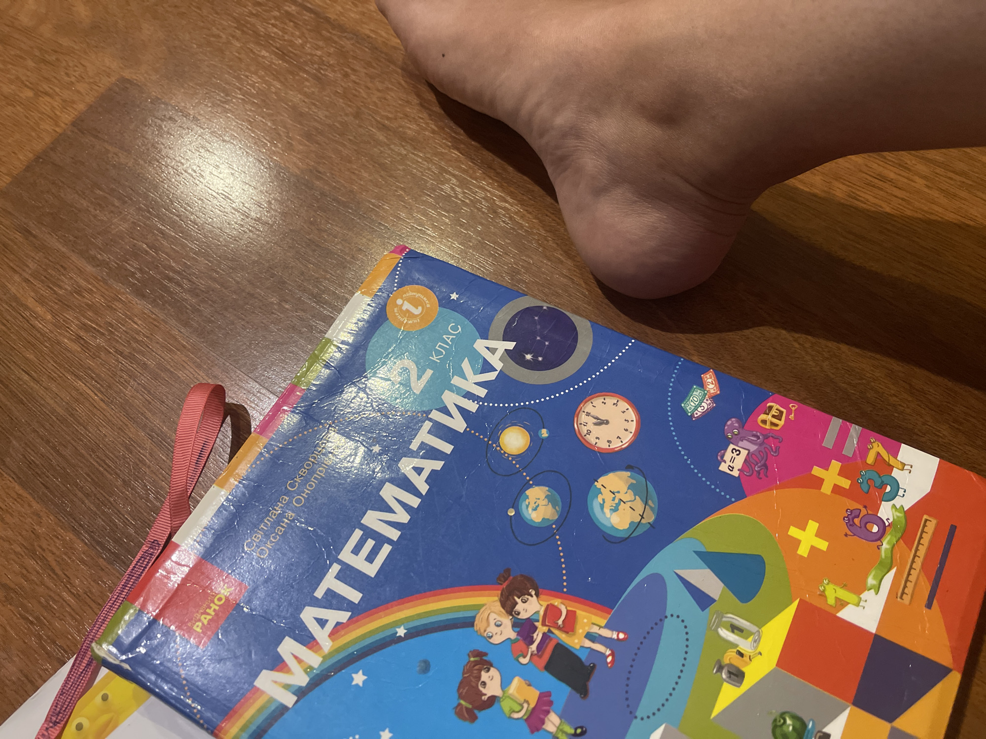 The image shows a child's mathematics textbook on a wooden floor, with a person's foot to the right. The textbook is blue and features colorful illustrations of children, planets, a rainbow, and mathematical symbols. The title "МАТЕМАТИКА" (Mathematics) is prominently displayed, along with "2 КЛАС" (2nd Grade). The textbook has a red ribbon bookmark. The wooden floor appears to be a laminate or hardwood, with visible grain and reflections from lighting. The foot belongs to an adult and is positioned in the lower right portion of the frame. The overall scene suggests a home environment. There is no indication of the location being in Poltava, Ukraine.