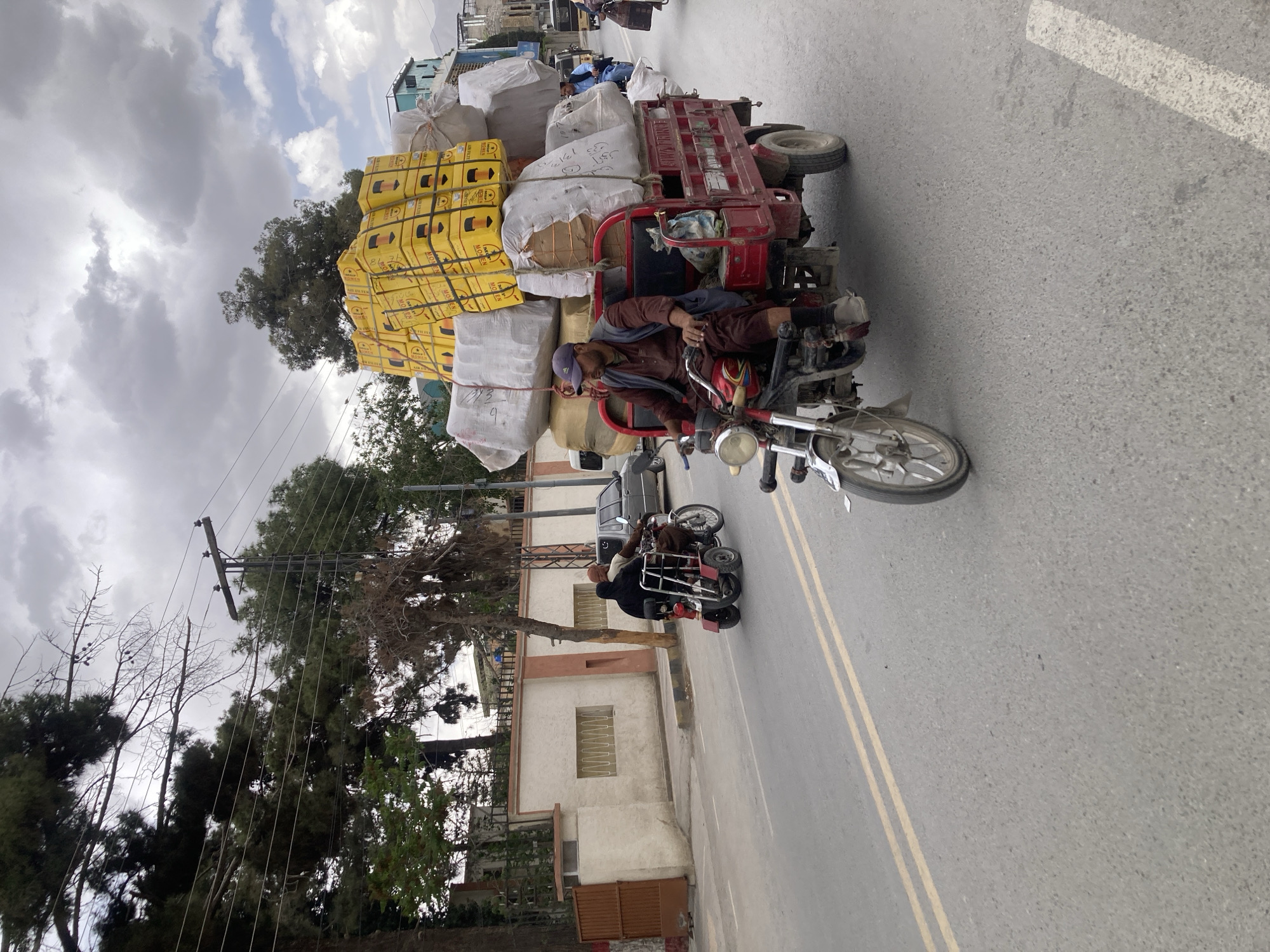 The image depicts a three-wheeled motorcycle heavily laden with goods traveling on a paved road in Quetta City Tehsil, Pakistan. The vehicle is loaded with yellow boxed goods and white sacks, towering over the motorcycle. The driver, wearing a dark brown jacket and blue cap, is seated on the motorcycle. Another individual is visible on a second motorcycle following behind. The background includes buildings, trees, power lines, and an overcast sky. The road is marked with parallel yellow lines.