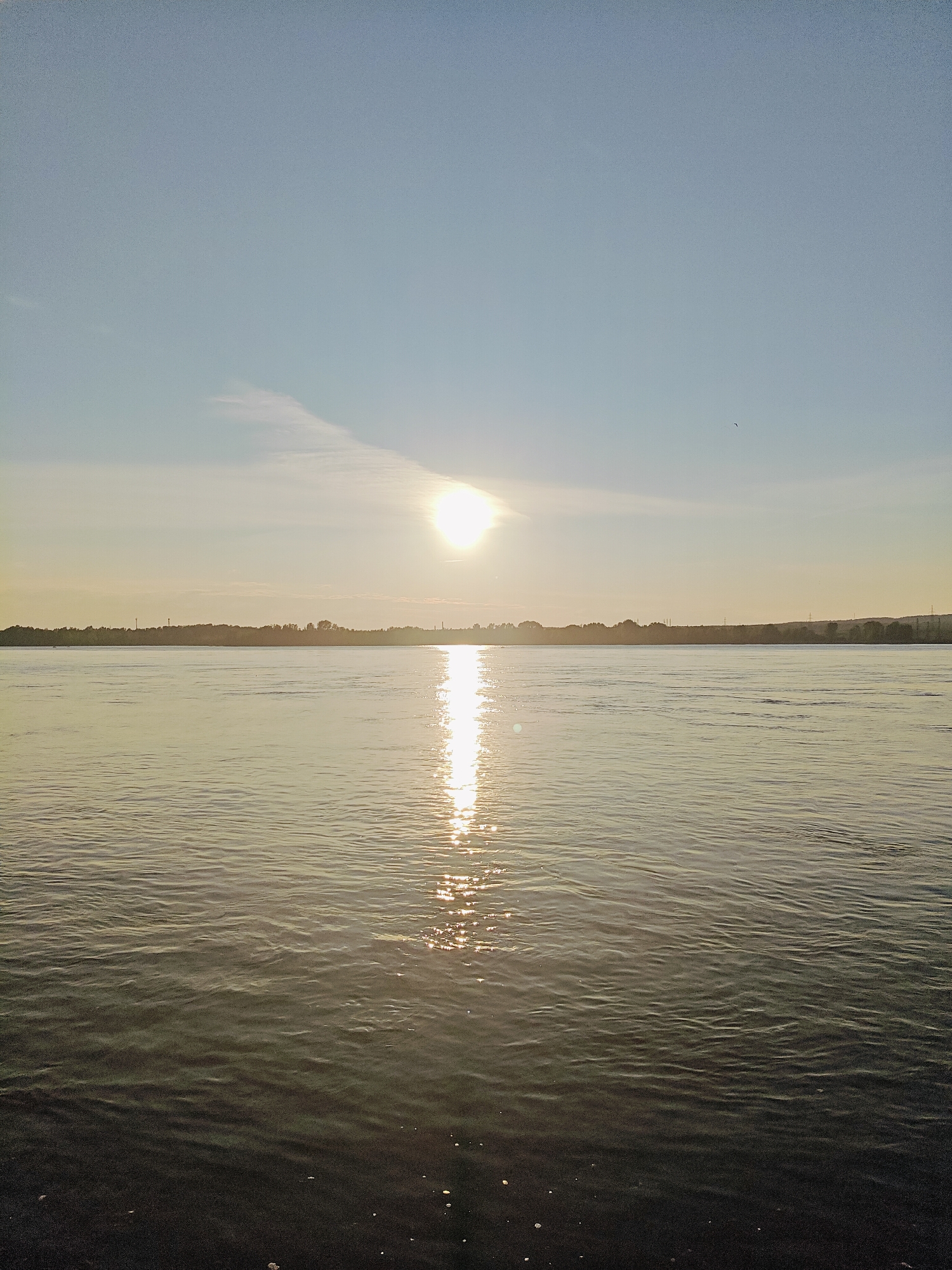 The image captures a serene sunset over a wide river, with the sun's bright reflection shimmering on the water's surface. A distant, tree-lined shore with a few structures is visible on the horizon. The sky is a soft blue, transitioning to a lighter, warmer hue near the horizon, with a few wispy clouds. The water has a gentle ripple, creating a textured pattern that catches the golden light. The overall mood is peaceful and tranquil, characteristic of a calm evening. The time of day is sunset, and the weather appears clear and calm. The location is implied to be near Chaikovsky, Russia, a city situated on the Kama River, which aligns with the visual elements of a broad river and its banks. There is no visible text in the image.