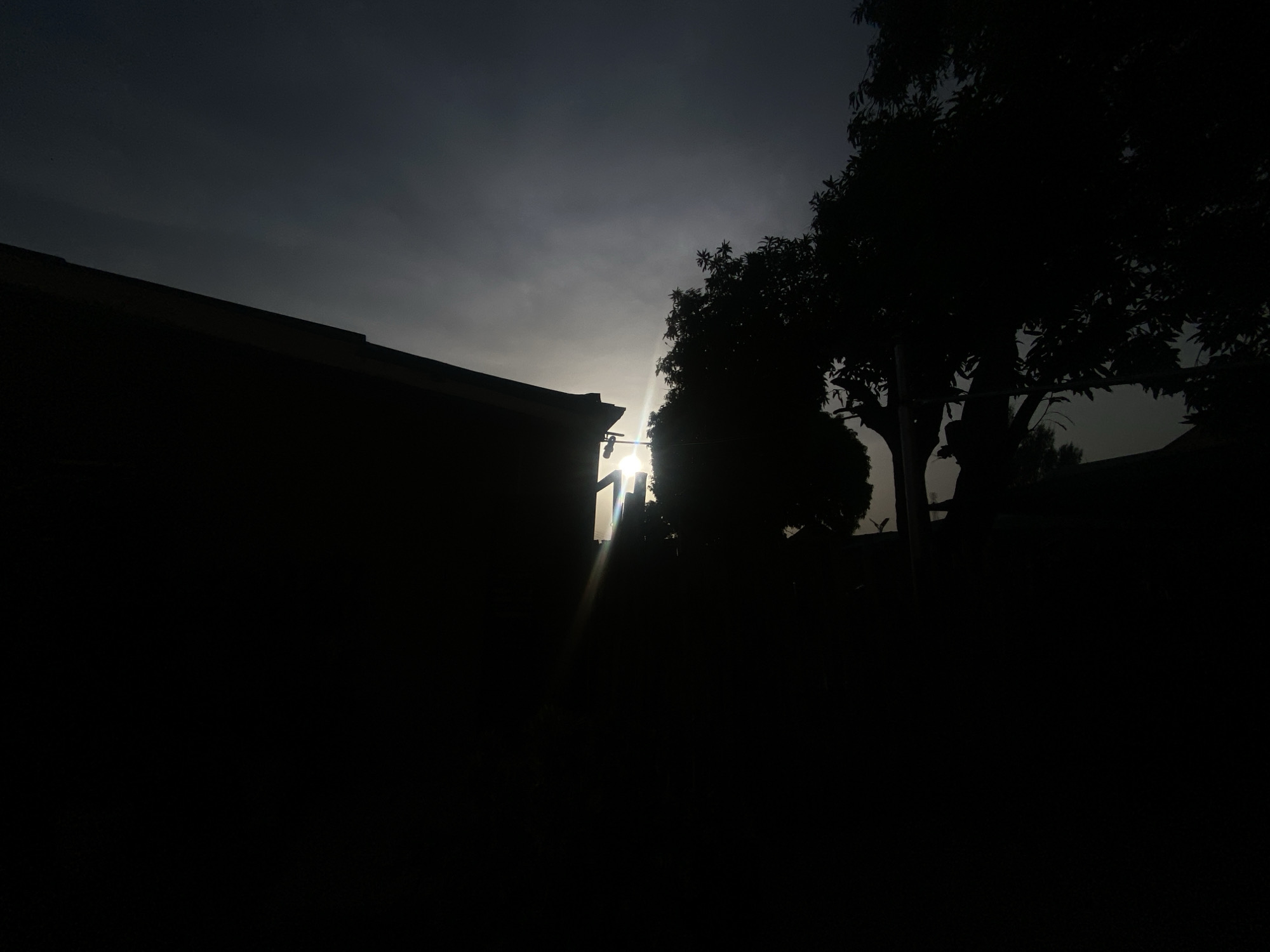 The image shows a silhouetted scene in Barden Yana, Nigeria. A building's roofline is visible on the left. A tree stands to the right of the building, and its foliage is also silhouetted. Light appears to be shining from behind the tree, creating a lens flare effect. The overall scene is dark, with most details obscured by shadow.