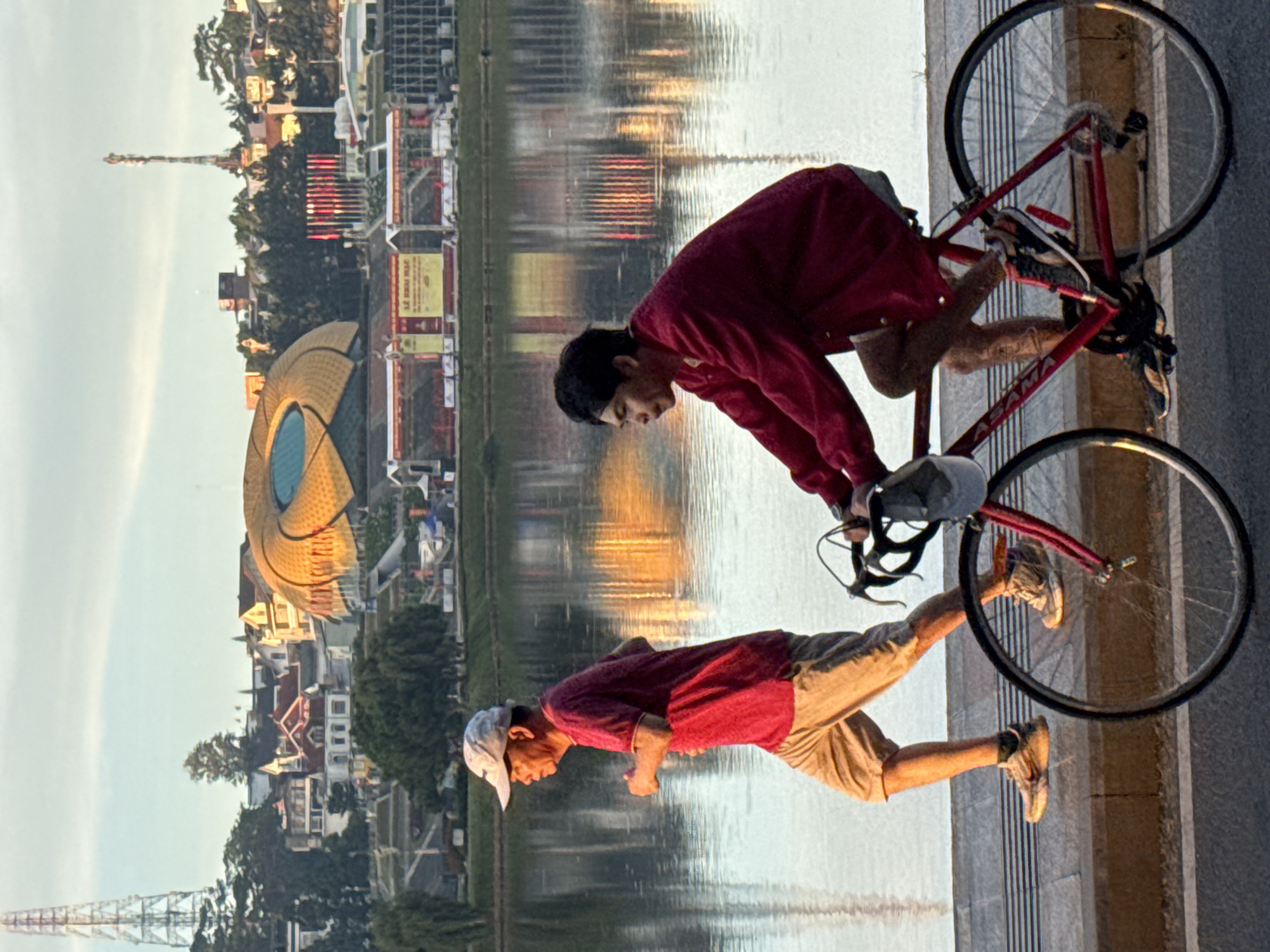 The image depicts a waterfront scene in Da Lat, Vietnam, at sunrise. In the foreground, an older man in a red shirt and beige shorts jogs along a paved path beside a calm lake. To his right, a younger man rides a red bicycle, also wearing a red long-sleeved shirt. The lake reflects the golden hues of the rising sun and the surrounding cityscape. In the background, a cluster of buildings, including a distinctive stadium with a golden, petal-like roof, is visible on a hill. Transmission towers and flags also dot the skyline. The overall atmosphere is tranquil and suggests a popular spot for morning exercise.