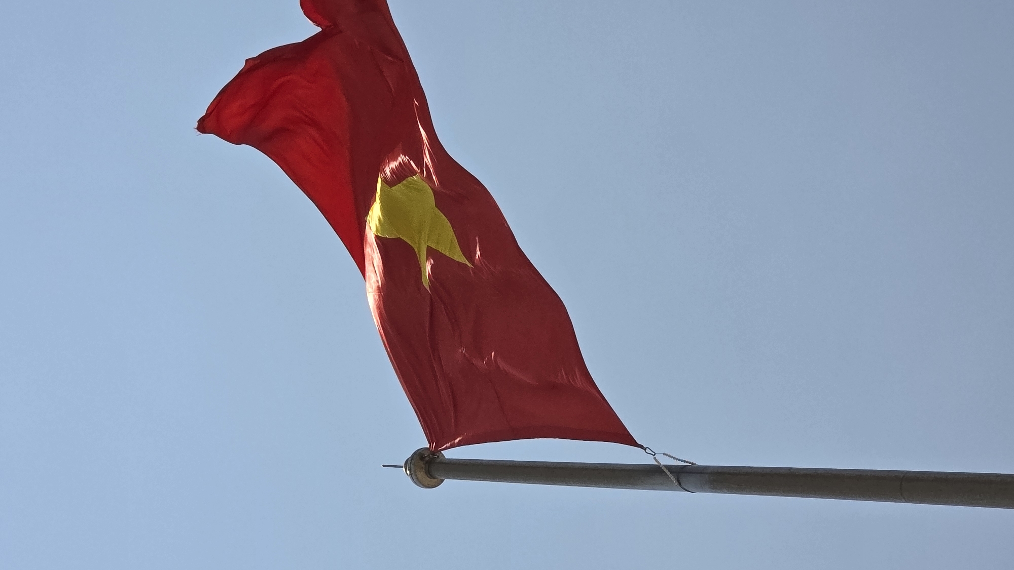 The image features the flag of Vietnam waving in the wind against a clear blue sky. The flag is predominantly red with a central yellow star. It is attached to a grey flagpole. The image likely captures a scene in Nha Trang, Vietnam, given the flag's national origin. There are no people visible in the frame.