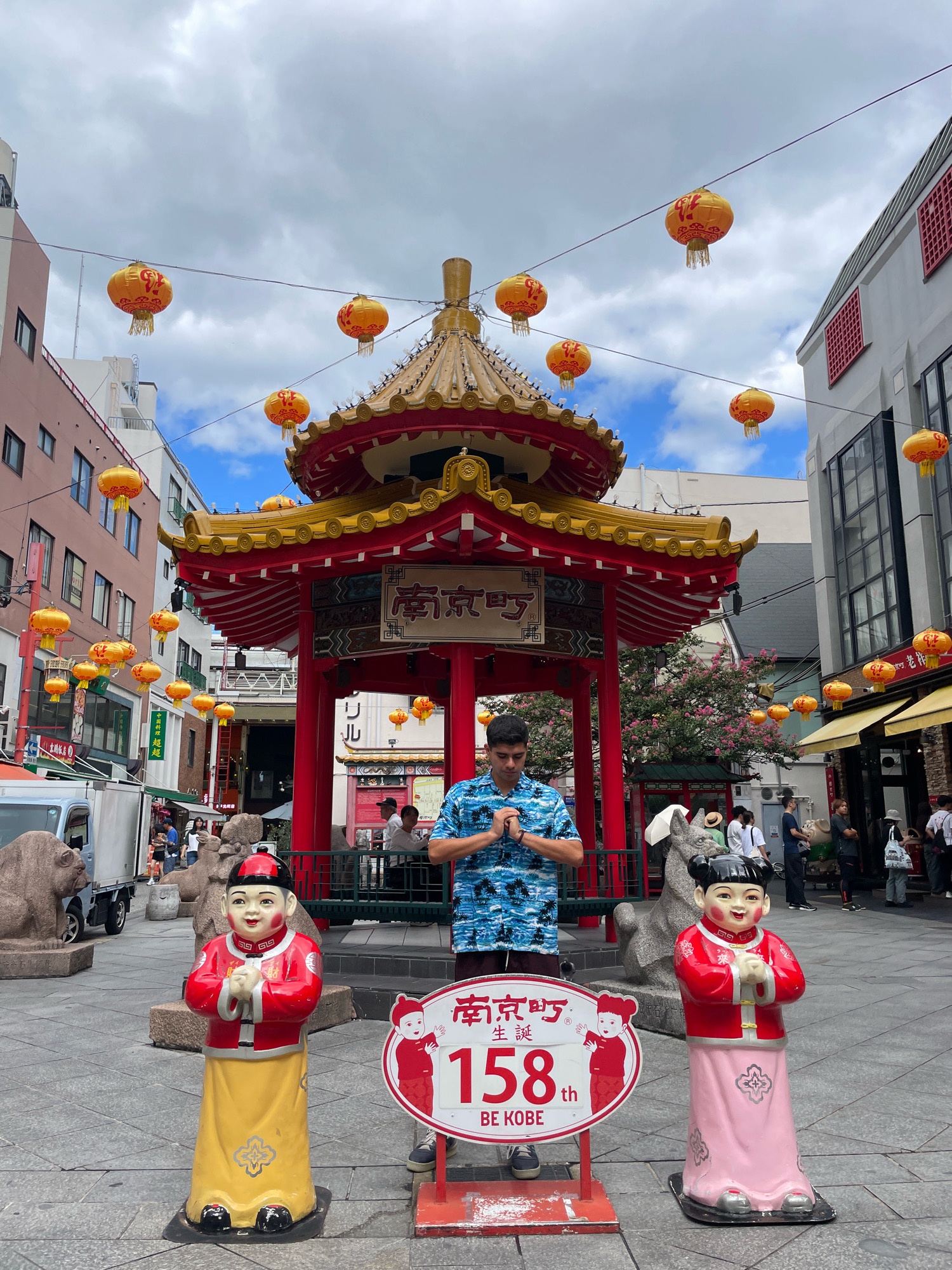 The image depicts a man standing in front of a traditional red and gold Chinese pagoda, with his hands pressed together in a gesture of prayer. The man is wearing a blue and white patterned short-sleeved shirt. In the foreground, there are two life-sized statues of children in traditional Chinese attire. The statue on the left is a boy wearing a red jacket and yellow skirt, while the statue on the right is a girl wearing a red jacket and pink skirt. Both statues are in a similar praying posture. A sign between the statues displays the number "158" and the text "BE KOBE". The scene appears to be in a public square, possibly a Chinatown, decorated with numerous yellow and orange lanterns hanging from wires overhead. Buildings with traditional Chinese architectural elements are visible in the background, as are other people walking around. The sky is cloudy. The location is identifiable as Nankinmachi, Kobe's Chinatown, given the sign and the style of architecture.