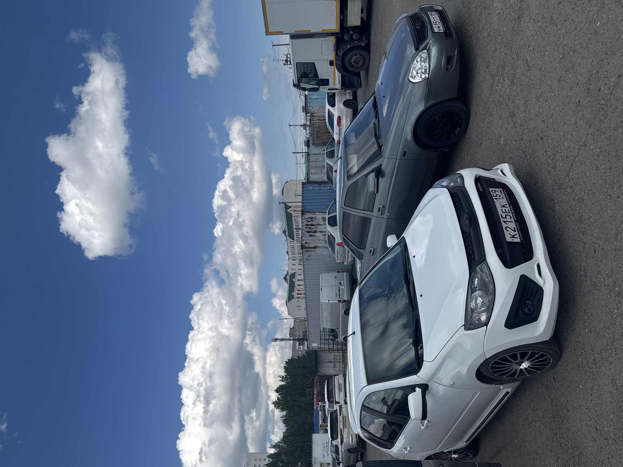 The image depicts a parking lot with several cars, including a white Lada Granta and a gray Lada Priora, against a backdrop of buildings and a blue sky with clouds. The license plate K215EK 159 on the white car indicates that the location is in Perm, Russia. Additional vehicles, including vans and a truck, are parked in the background. The setting appears to be a commercial or industrial area.