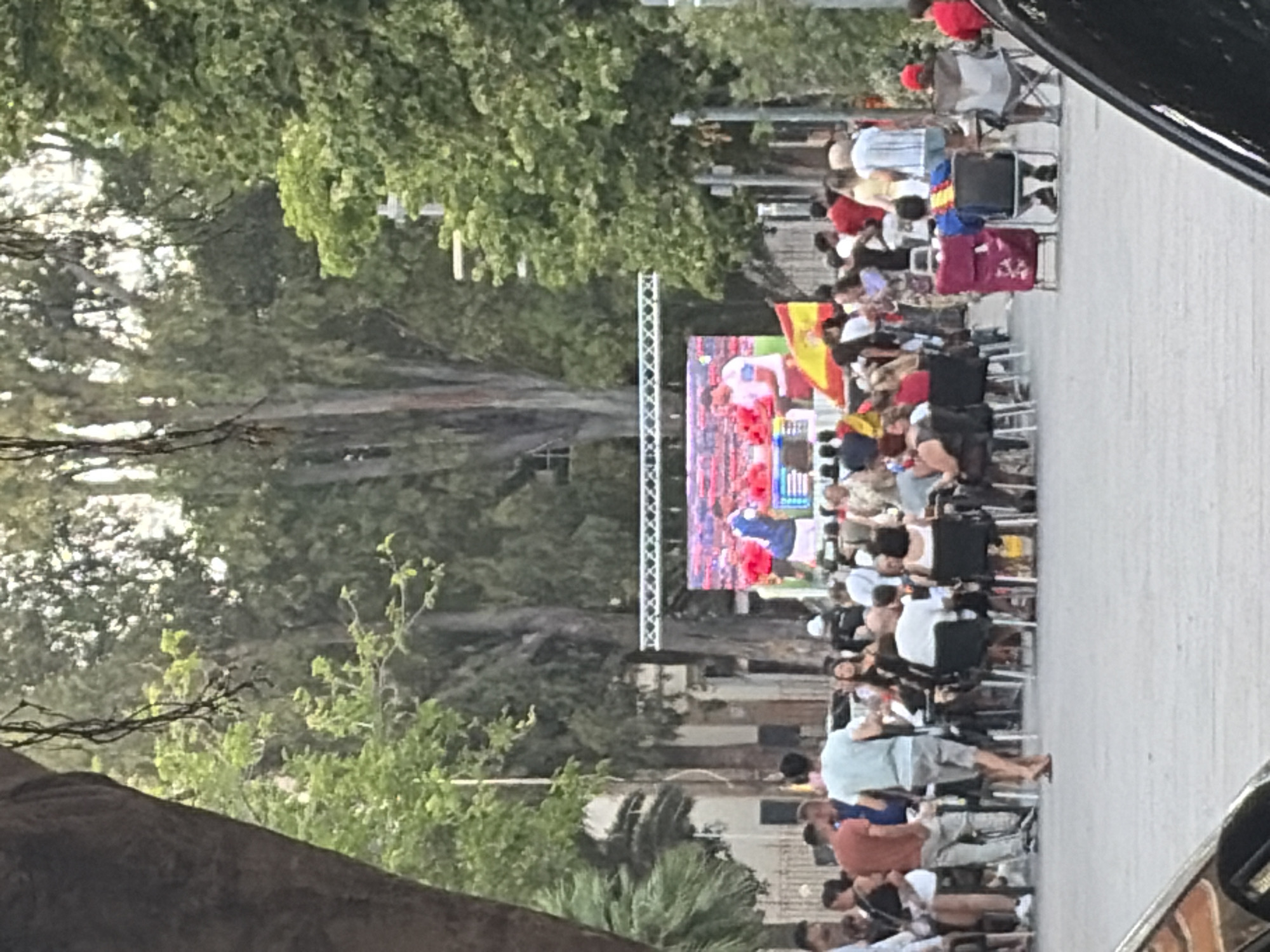 The image depicts an outdoor viewing of a soccer match on a large screen, possibly at a public event or in a plaza in Sagunto/Sagunt, Spain. A crowd of people is gathered, seated at tables or standing, facing the screen. Some individuals are wearing shirts with the Spanish flag colors, and Spanish flags are visible in the foreground, suggesting support for the Spanish national team. The screen displays two figures in soccer uniforms, one in blue and white and another in red and white, possibly celebrating a goal. Trees and buildings form the backdrop. The overall atmosphere appears to be festive and communal.