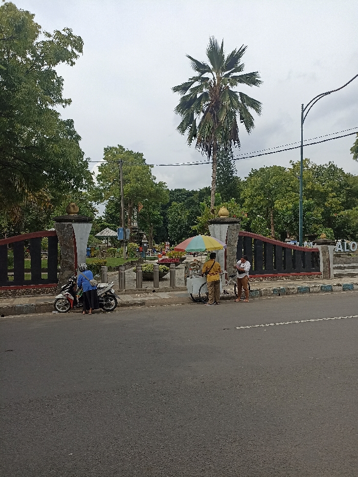 The image captures an outdoor scene in Tulungagung, Indonesia, under an overcast sky during daytime. A paved road occupies the foreground, bordered by a curb and a sidewalk.
On the left side of the frame, two individuals are standing next to a white and red motor scooter parked at the curb. One person, wearing a blue top and black pants or skirt, is positioned near the scooter, appearing to interact with it or prepare for departure.
To the right, a street vendor operates a white pushcart topped with a colorful striped umbrella (featuring shades of red, yellow, light blue, and green). The vendor, dressed in a yellow shirt and brown pants, stands behind the cart. A customer, wearing a light-colored top and brown pants, stands in front of the cart, suggesting a transaction is underway.
Behind the street and sidewalk is the entrance to what appears to be a park or public garden. The entrance is marked by decorative pillars and a dark fence-like structure. Lush green trees and dense foliage fill the background, with a very tall palm tree prominently standing near the center-right. Further inside the park, more trees and some smaller structures, possibly a gazebo, can be seen. A street light pole is visible on the far right. Part of a sign on the park's entrance wall is visible, displaying the letters "ALO", which likely forms part of "Alun-alun", a common term for a town square or public park in Indonesia. The scene depicts typical daily activities beside a public green space.