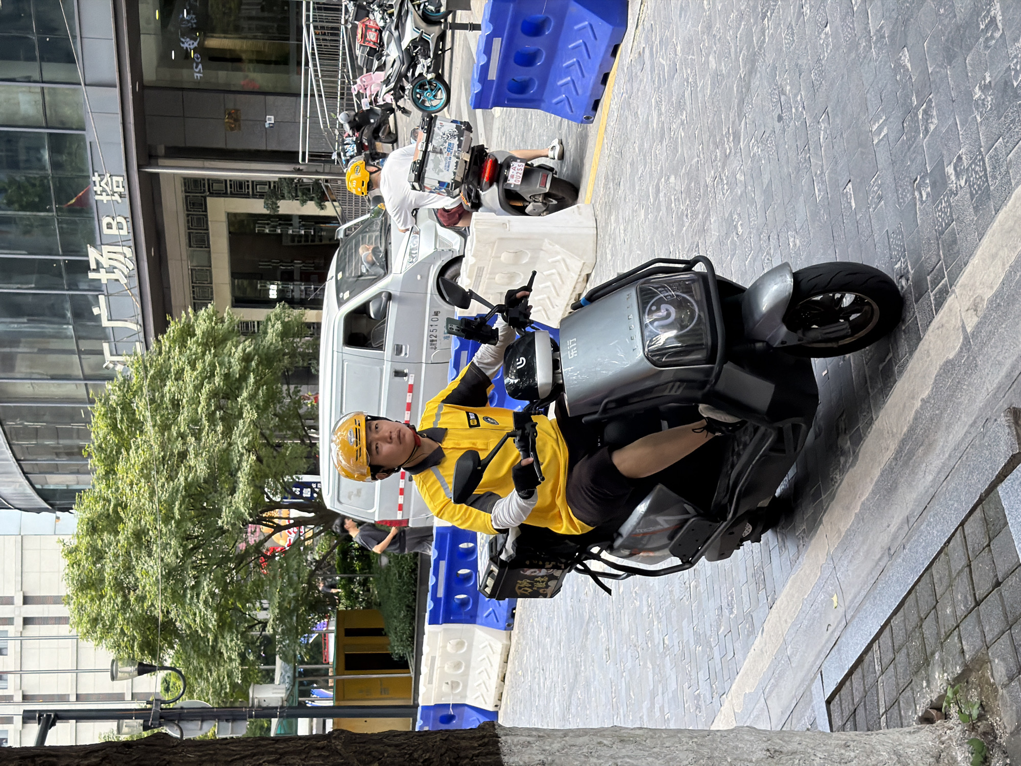 In Yuzhong District, China, a male delivery person wearing a yellow shirt, black shorts, and a yellow helmet rides a silver electric scooter. The scooter has a black storage box on the back, adorned with colorful stickers. The delivery person is holding the handlebars and appears to be looking forward. The scene takes place on a city street with a cobblestone sidewalk. Blue and white temporary barriers line the street. In the background, a white van is parked, and various buildings and trees are visible.