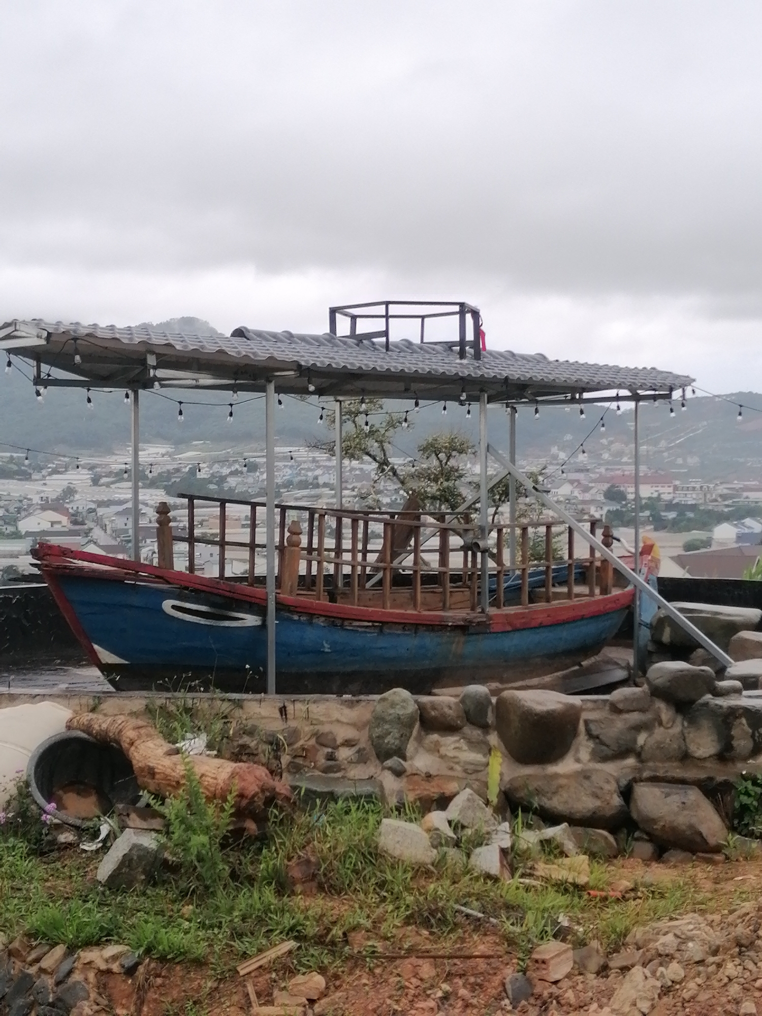 The image depicts an outdoor scene in Lạc Dương, Vietnam, featuring a modified boat serving as a shelter or seating area. The boat, painted blue with a red trim, is positioned under a weathered, gray tiled roof supported by metal poles. String lights are draped across the roof structure. The boat's interior is lined with wooden railings. In the background, a hazy view of a city or town with buildings nestled on rolling hills under an overcast sky is visible. In the foreground, the area around the boat is rustic, with a stone wall, rocks, and patches of grass and dirt. A large, dark planter with some greenery and a piece of gnarled wood are also present in the foreground. There are no people visible in the image.