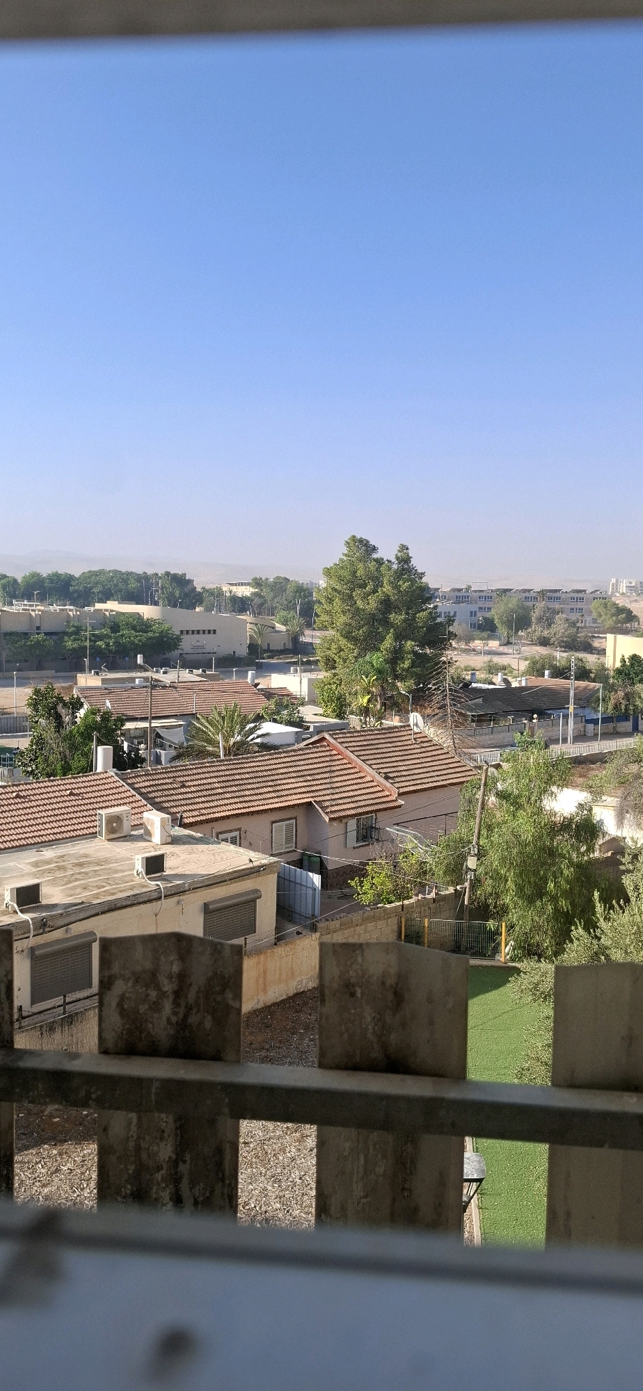 The image captures an aerial view of a residential area in Yerocham, Israel, under a clear blue sky. In the foreground, a concrete structure with metal railings is visible, partially obscuring the view below. The scene reveals a cluster of houses with red-tiled roofs and light-colored walls, typical of Israeli architecture. Several of these houses have air conditioning units mounted on their roofs or walls.
Beyond the immediate houses, there is a mixture of vegetation, including trees and shrubs, interspersed with more buildings. Some of these buildings appear to be larger, possibly institutional or commercial, with beige and white facades. A prominent, large tree stands out in the middle ground, casting a shadow.
Further in the distance, the landscape transitions into a more arid environment, with low hills visible on the horizon, characteristic of the Negev desert region where Yerocham is located. The overall impression is one of a sun-drenched, semi-arid urban environment. No people or active events are depicted in the image.