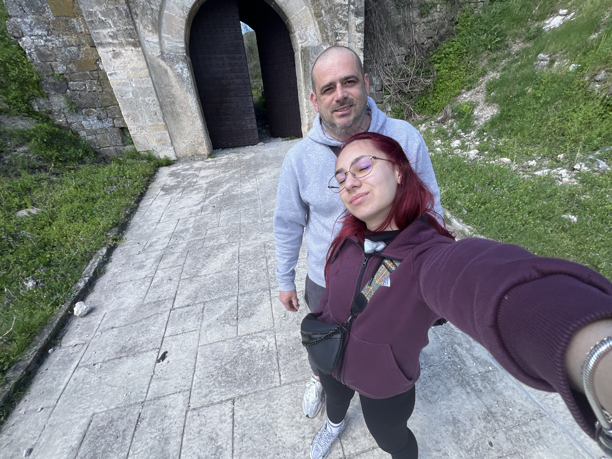 A wide-angle, eye-level shot depicts a man and a woman posing for a selfie in front of a stone structure with a large arched doorway. The doorway features wooden doors that are partially open. The path leading to the doorway is paved with large stone slabs. The woman is in the foreground, holding the camera, wearing a maroon hooded jacket, black leggings, and white sneakers. She has red hair and glasses. A black crossbody bag is visible. The man stands slightly behind her, wearing a gray hoodie. The background includes green foliage on either side of the path. The location appears to be outdoors in the city of Nikopol, Bulgaria.