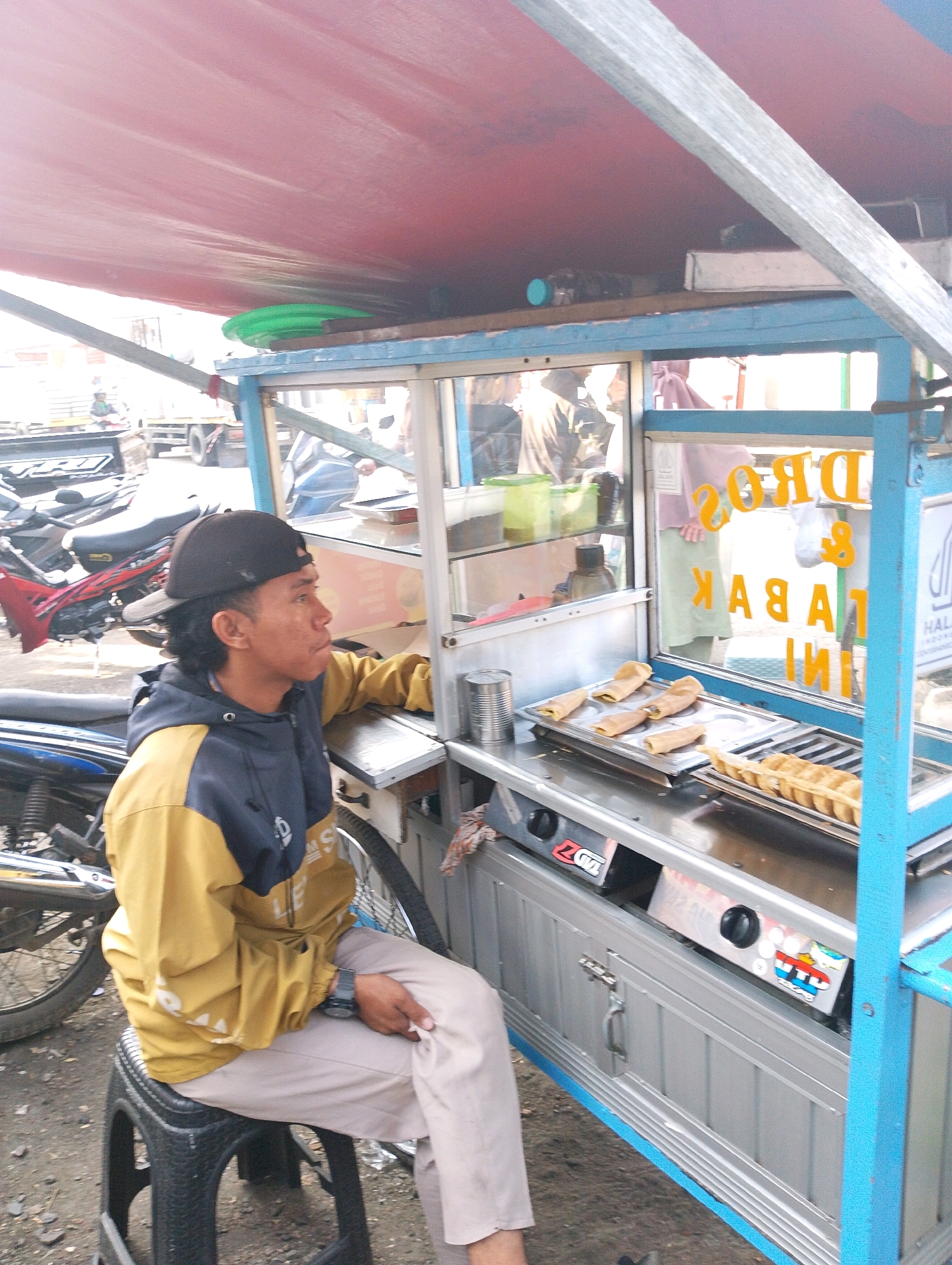 In Lembang, Indonesia, a street vendor prepares crepes on a mobile cart under a red awning. The cart features a glass display case and a cooking surface with two gas burners. A man wearing a yellow and dark blue hooded jacket and a black baseball cap sits on a black stool, looking at the crepes. The background shows parked motorcycles and a truck. The cart's signage is visible, with the words "DROS & TABAK" in orange lettering.
