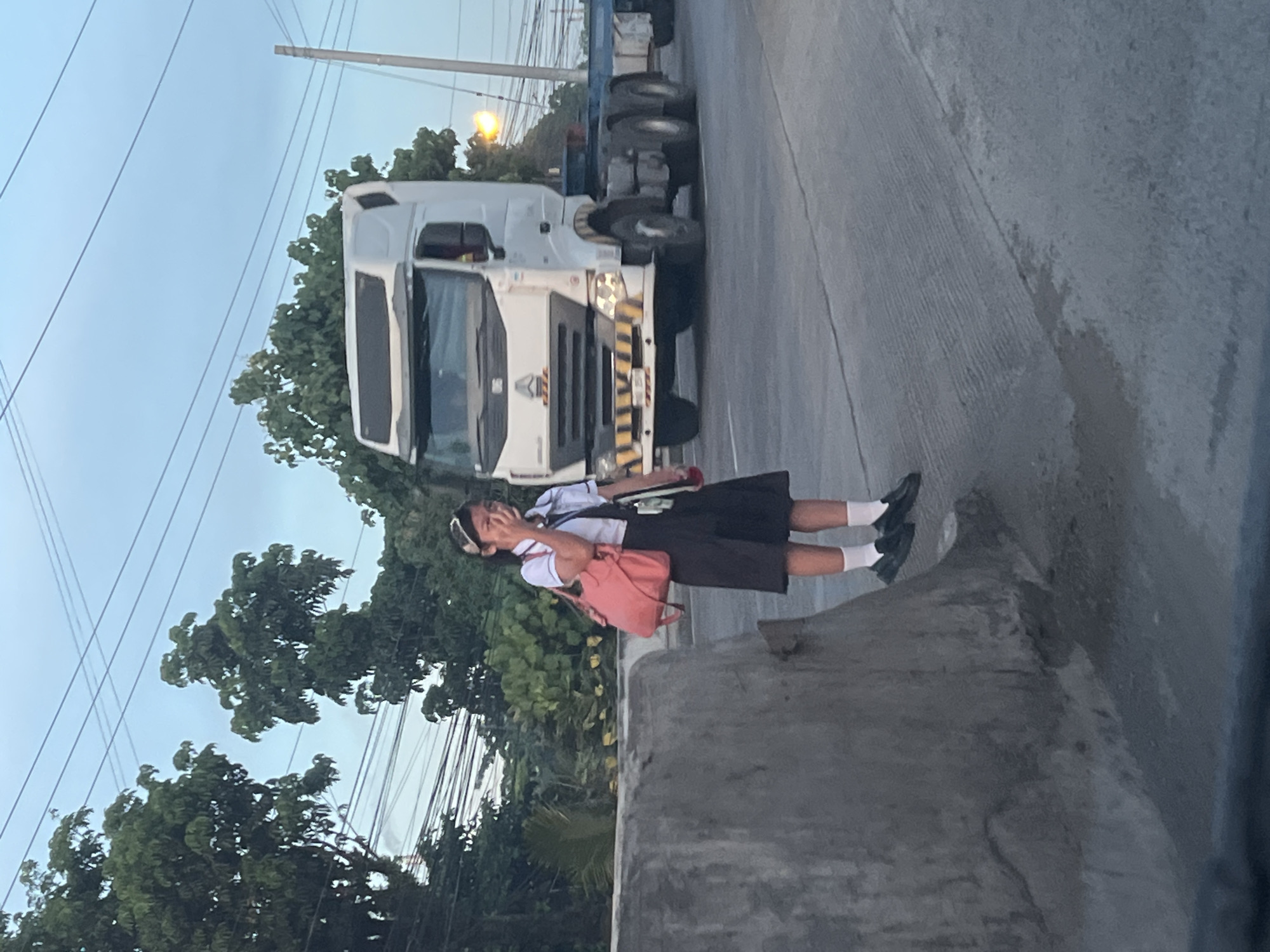 A young person wearing a school uniform, complete with a white polo shirt, dark skirt, white socks, and black shoes, stands by a concrete barrier. They are holding a pink bag on their left shoulder and appear to be crying, with their right hand covering their mouth and eyes. Behind them, a white semi-trailer truck is driving on a road. The scene is set against a backdrop of trees and overhead electrical wires, indicative of an urban environment. The lighting suggests it is either early morning or late afternoon. This scene is likely located in Davao City, Philippines, given the presence of the truck and the commonality of school uniforms in the region.
