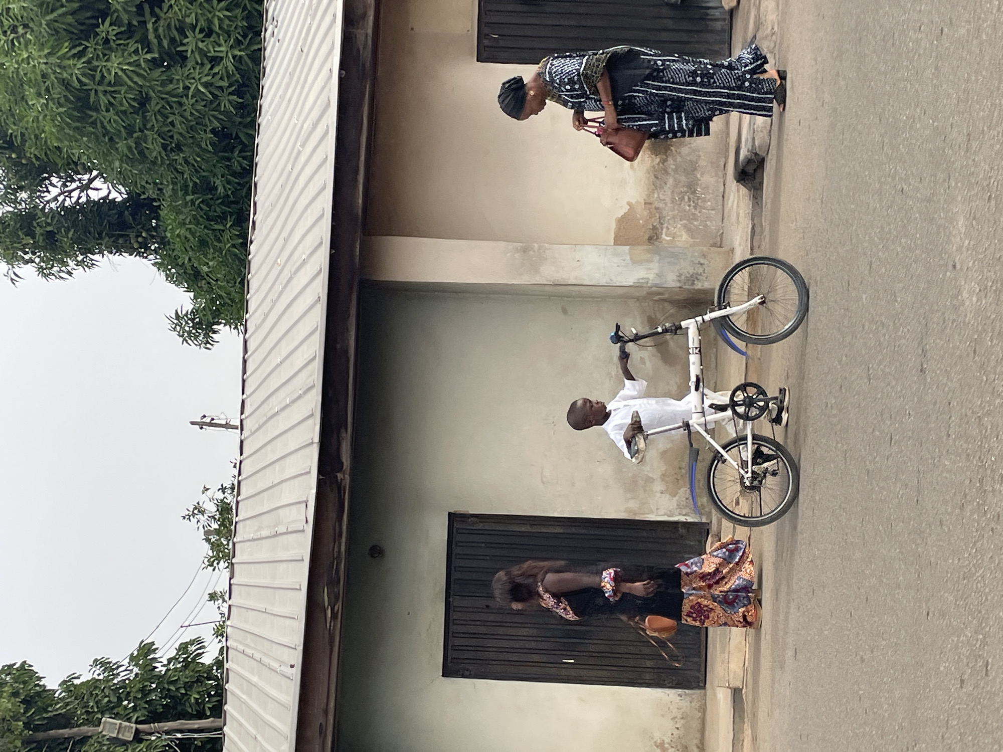 The scene is set on a paved road in Jos, Nigeria, beside a single-story building with a white facade.
An elderly woman in a patterned dress stands near the building corner, looking down.
A young person dressed in white stands next to a white bicycle, holding the handlebars. Another woman is standing in front of a closed black door.
A tree with dense green foliage occupies the left side of the image. Utility wires stretch across the sky.