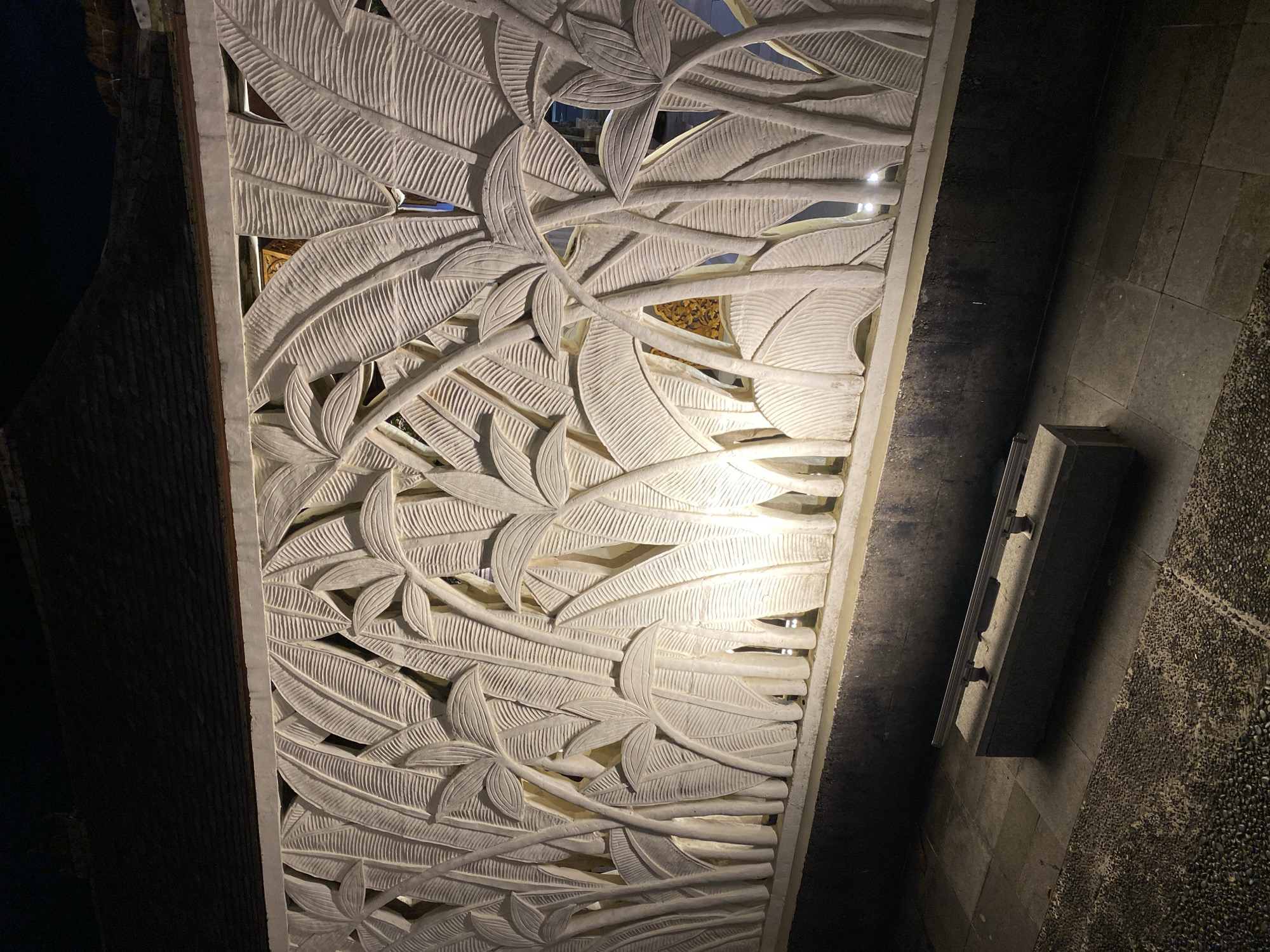 The image shows a close-up of a textured white wall carved with tropical foliage, likely palm or banana leaves, at night. The carving is illuminated from within, casting shadows and highlighting the intricate details of the leaves and stems. The wall appears to be part of an outdoor setting, possibly a garden or courtyard. In the foreground, a stone bench with a metal railing is visible on a paved area. The ground is covered with large square tiles and smaller pebbles. The top of the image reveals a dark roof structure, suggesting a building adjacent to the wall. The scene is set in Kuta Selatan, Indonesia.