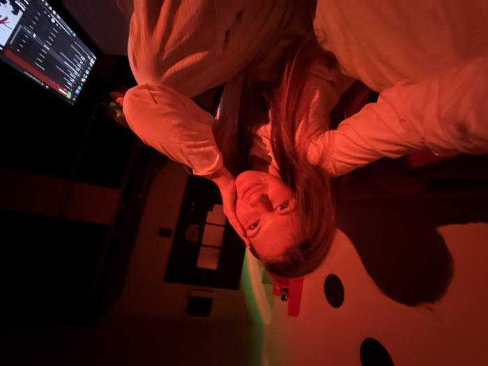 An indoor scene is depicted, illuminated predominantly by red light with a faint green tint in the upper left. A female subject is positioned centrally, taking a selfie, extending her right arm towards the camera. She has long, light brown hair and a slight smile. Her left hand rests on her left cheek. She wears a light-colored, possibly beige, collared long-sleeved shirt and matching trousers. The background to the left features a white wall with a large, stylized black and white design, possibly resembling a cartoon face or abstract symbol, upon which a red shadow is cast. To the right, a dark, closed door with multiple rectangular glass panes is visible. Further to the right, a laptop screen displays a user interface consistent with a music streaming application, such as Spotify. The specific location within the city of Vozdovac Urban Municipality, Serbia, is not discernible from the visual evidence.