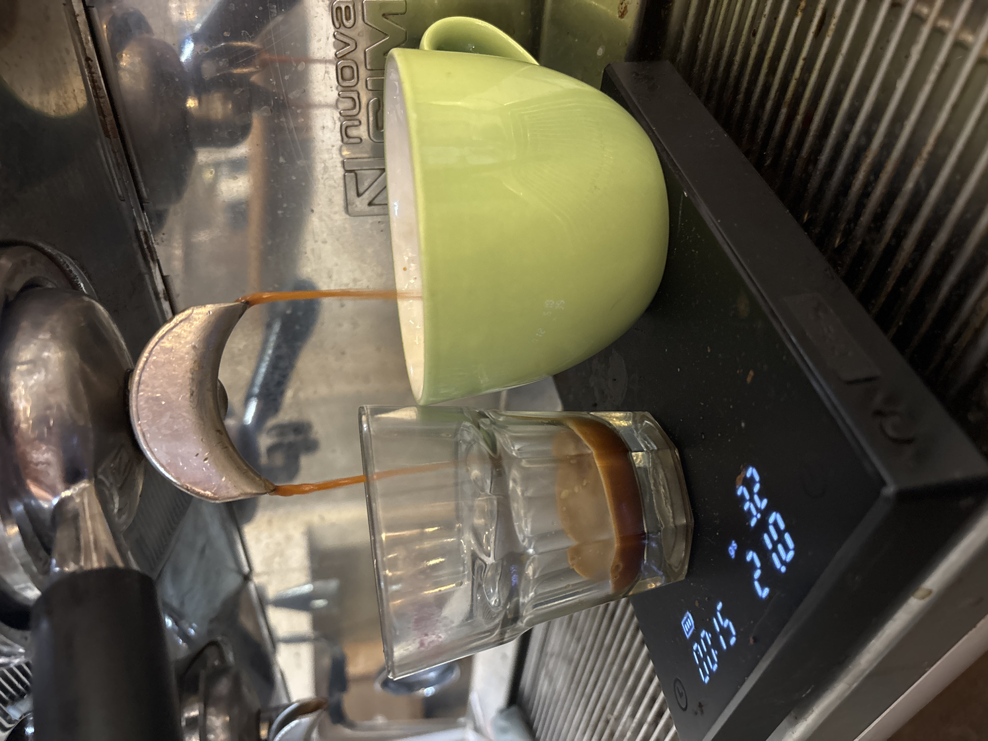 An overhead, rotated close-up shot captures the precise extraction of espresso from a coffee machine, likely in a cafe or kitchen setting in Buenos Aires, Argentina. Two thin, dark amber streams of espresso are seen flowing downwards from the double spouts of a metal portafilter. One stream enters a clear, faceted glass, where a rich, light brown crema layer is visibly forming on top of the dark liquid.
The glass rests on a black digital scale, which displays measurements in blue-white digits: '00:15' (indicating 15 seconds of extraction), '27.0' (likely 27.0 grams or milliliters of liquid), and '32' (possibly a different measurement unit or target weight). Beside the glass, a light green ceramic coffee cup is inverted, resting on the machine's surface, with the second stream of espresso flowing into or past it. The background reveals parts of a well-used espresso machine, including a shiny, scratched metal surface with the partial brand name "nuova" visible on a silver plate. Below the scale and glass, a ribbed metal grate, part of the drip tray, is visible. The scene emphasizes the meticulous process of coffee making.
