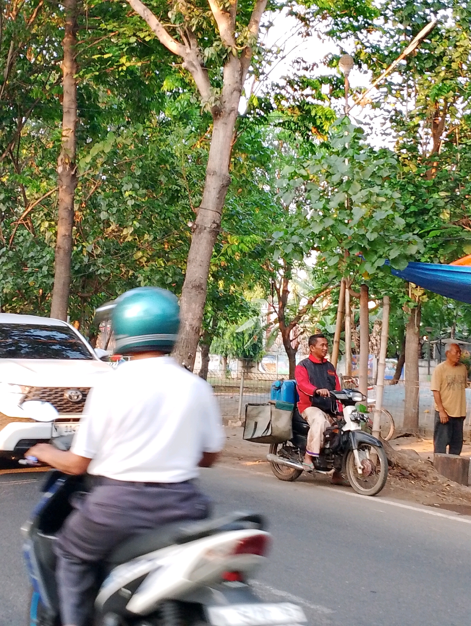 This is a street-level, medium-shot photograph taken outdoors in Probolinggo, Indonesia, likely during daylight hours with moderate lighting. The scene depicts a road with several individuals and vehicles.
In the foreground, on the left, a person is riding a motorcycle and is heavily blurred due to motion. They are wearing a white t-shirt, dark pants, and a teal motorcycle helmet. A white car is partially visible to the left of this motorcycle rider, parked or moving slowly.
Further down the road, on the right side, a man is sitting on a black and white motorcycle. He is wearing a red jacket with black accents and beige pants. The motorcycle has two large bags attached to the back. To the right of this man, another man stands on the sidewalk wearing a light brown t-shirt and dark pants.
The environment consists of a paved road, a dirt embankment, and a backdrop of numerous trees with dense green foliage. There are also what appear to be streetlights visible through the trees. Two large tarps, one blue and one orange, are visible hanging above the scene in the background. The overall impression is that of a busy street scene in a tropical or subtropical climate.