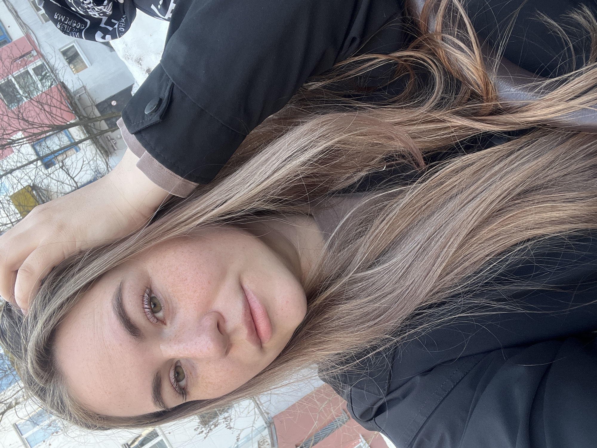 A selfie-style photograph depicts a young woman with light blonde hair and freckles on her face. She is wearing a black jacket. The background includes a building with red and white exterior walls and bare trees, indicative of winter. The setting appears to be urban, in the city of Chaikovsky, Russia. Snow is visible on the ground and on the building. The woman's hand is visible on the top of her head, suggesting she is holding her hair.