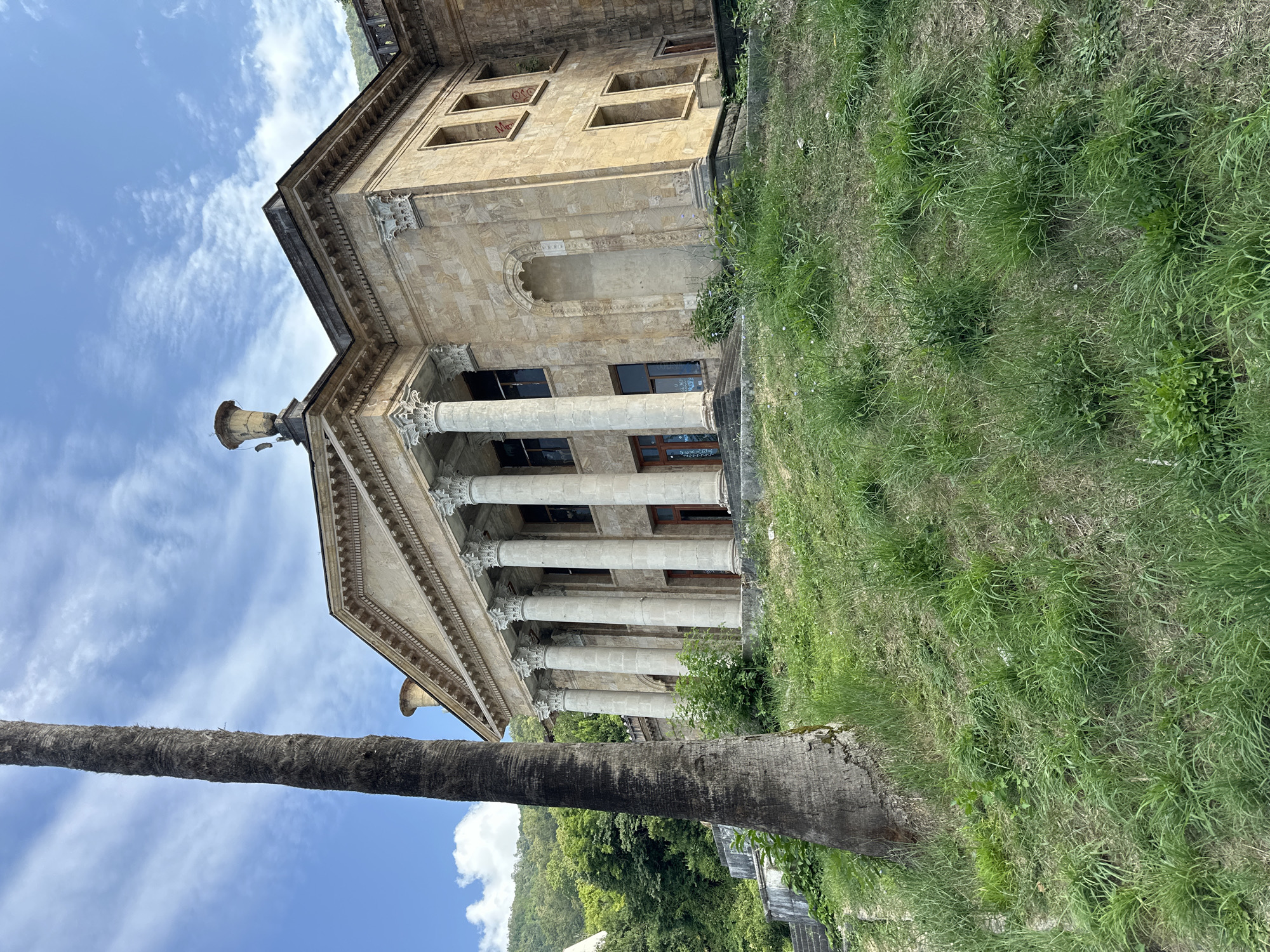 The image shows a large, old, stone building with a grand facade, featuring multiple tall columns and ornate architectural details. The building appears to be in a state of disrepair, with some windows boarded up or broken, and graffiti visible on the exterior walls. It is situated on a grassy slope, with a tall palm tree in the foreground to the left. Lush green vegetation and trees cover the hillsides in the background, under a bright blue sky with scattered clouds. The scene suggests an abandoned or neglected historical structure, possibly in a subtropical or Mediterranean climate. This building is the former sanatorium "Gagripsh" located in the city of Abaata, Abkhazia.