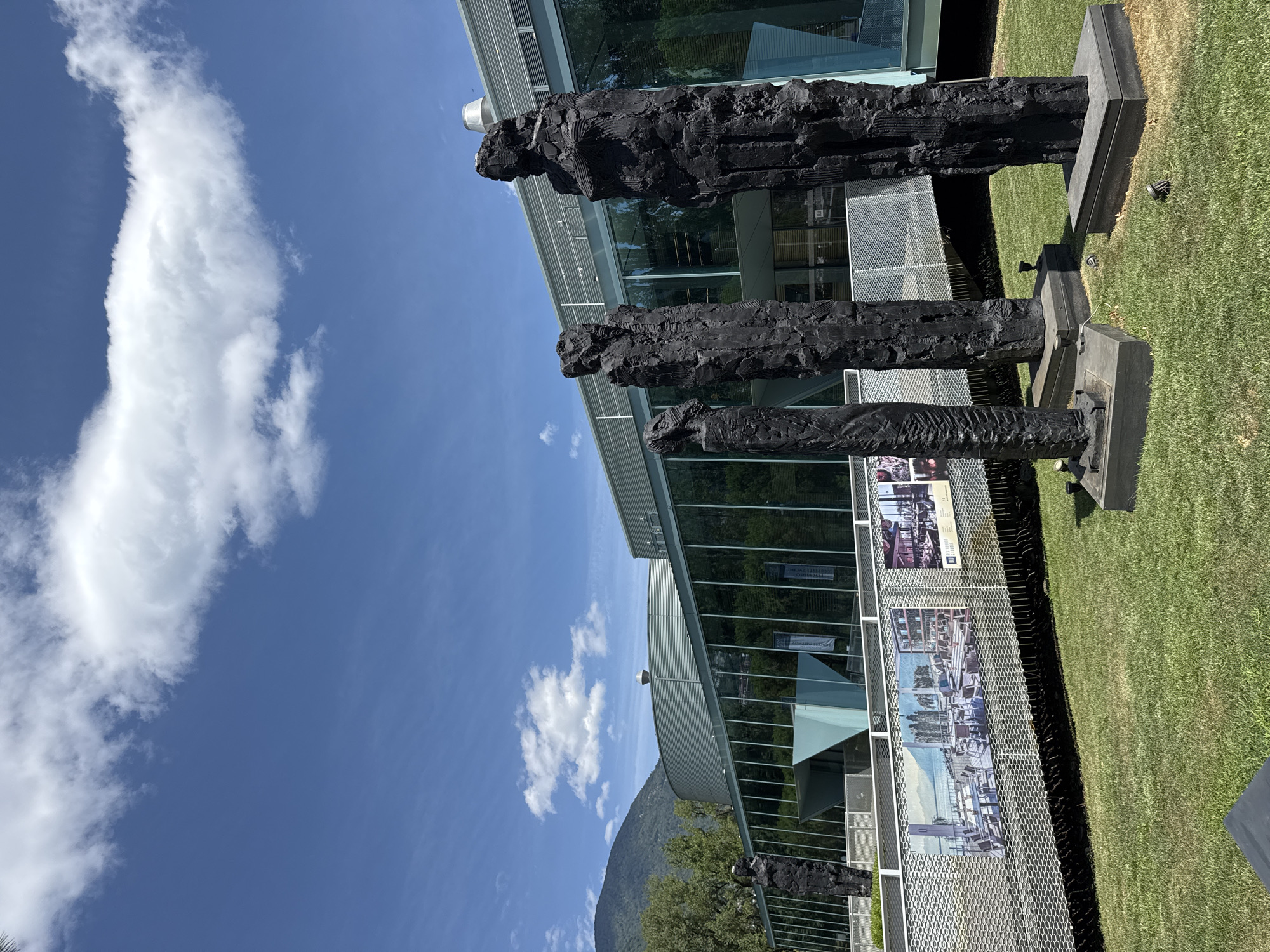 The image depicts the exterior of the Castello Visconteo in Locarno, Switzerland, on a clear day with scattered clouds. In the foreground, four tall, dark, abstract sculptures resembling human figures stand on square bases in a grassy area. Behind the sculptures, a contemporary building with a glass facade reflects the surrounding trees and sky. A metal walkway with a perforated surface runs horizontally across the mid-ground, displaying several large photographic panels or artworks. The building features a curved, metallic section and a geometric entrance. In the distance, a tree-covered mountain range is visible under the bright blue sky. The scene captures an outdoor public space adjacent to a historical building, showcasing a blend of modern architecture and art installations.