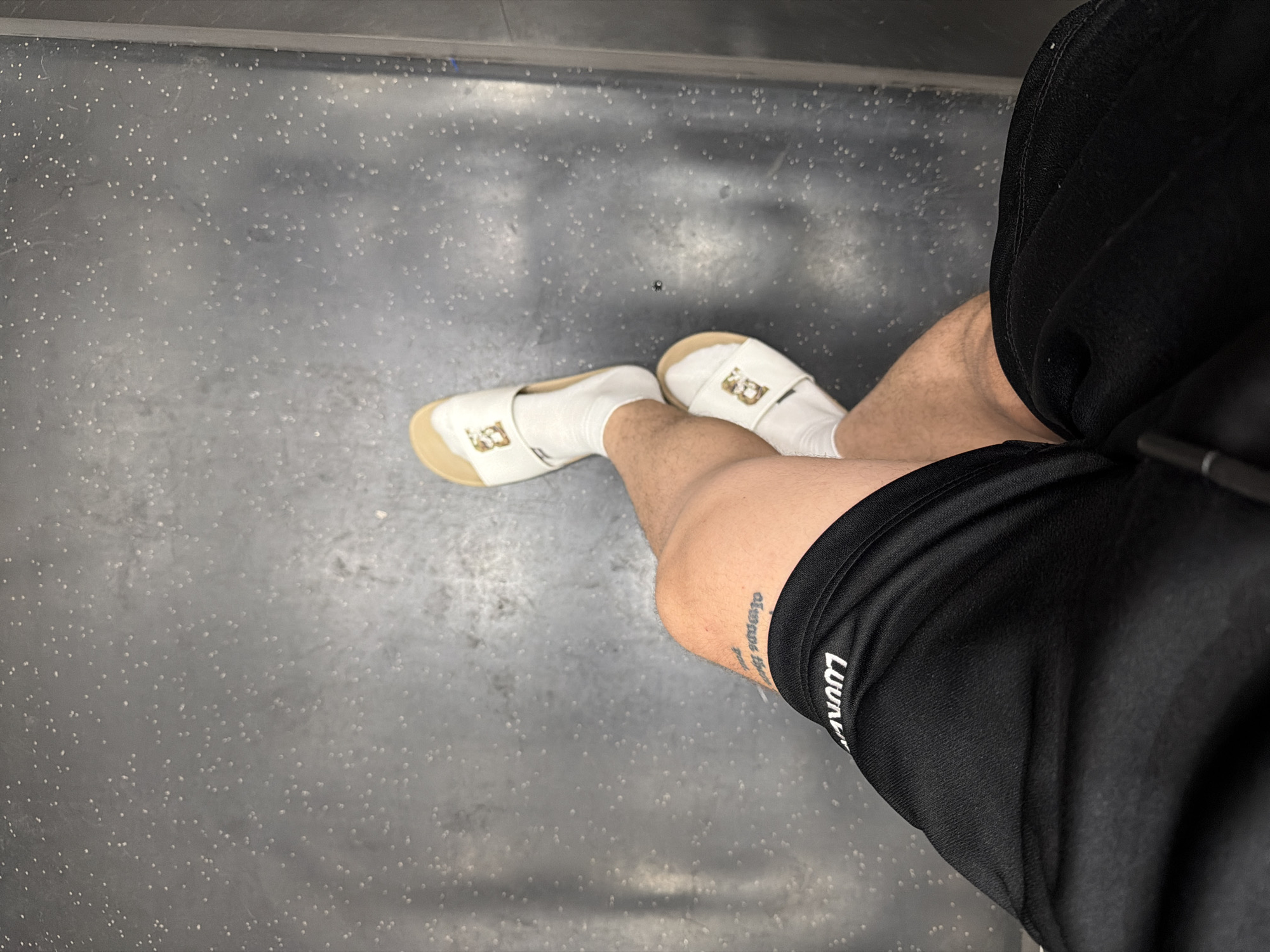 The image shows a person's legs and feet from a high-angle perspective, resting on a speckled gray floor. The person is wearing white socks and white slides with a gold buckle. Black athletic shorts are visible on the legs, with a white logo on the left leg that appears to read "LUUVIM". There is a tattoo on the left thigh, partially obscured by the shorts, which seems to contain cursive text. The floor appears to be a hard, textured surface, possibly a gym floor or a public transit area. The lighting is overhead and even. There are no other people or objects visible in the immediate vicinity. The image does not contain any discernible visual cues that would indicate the location as Bangkok, Thailand.