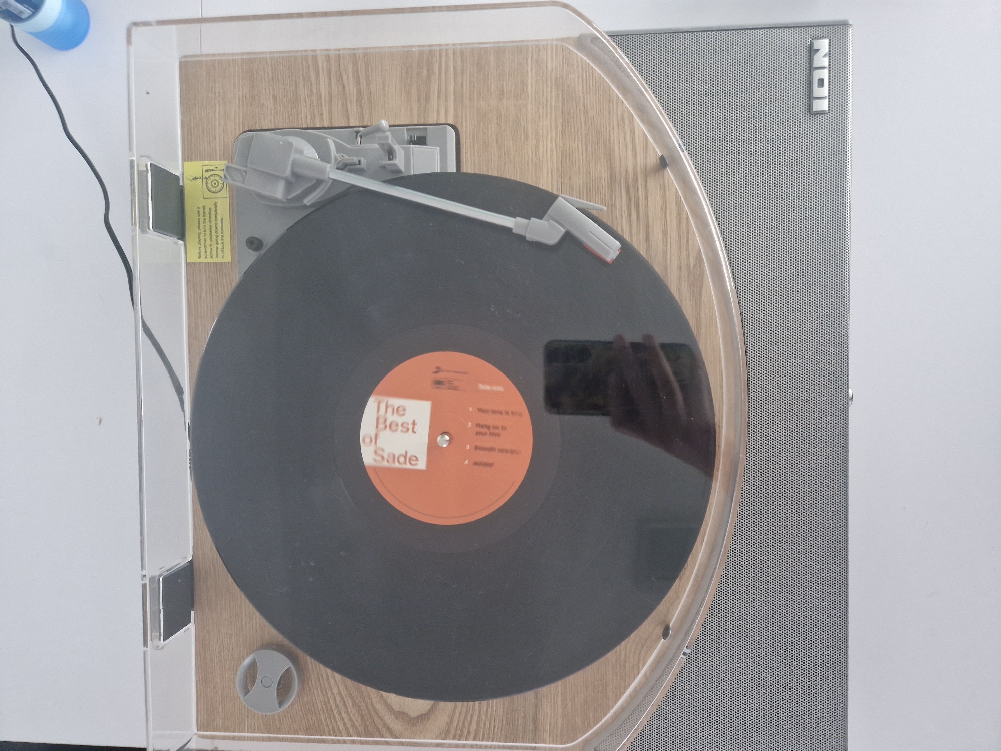 Here is a description of the image:
The image shows a turntable with a record on it. The record is "The Best of Sade." The turntable is an ION model and has a clear plastic dust cover that is open. A label on the left side gives instructions for unlocking the turntable before playing. The turntable sits on a light-colored surface. A hand holding a smartphone is reflected on the vinyl record.