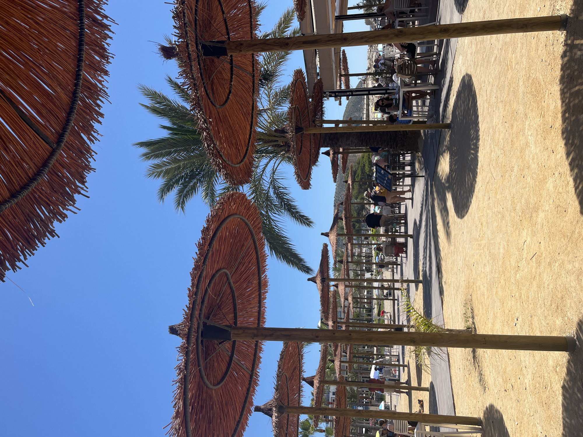 The image shows a beach area, likely a resort or restaurant, in Teulada, Spain. The scene is captured from a low angle, looking upwards through several rows of thatched beach umbrellas with wooden poles. A palm tree stands amidst the umbrellas. Tables and chairs are visible underneath the umbrellas, with people seated and dining. The ground is sandy. The sky is a clear blue. The umbrellas cast shadows on the ground.