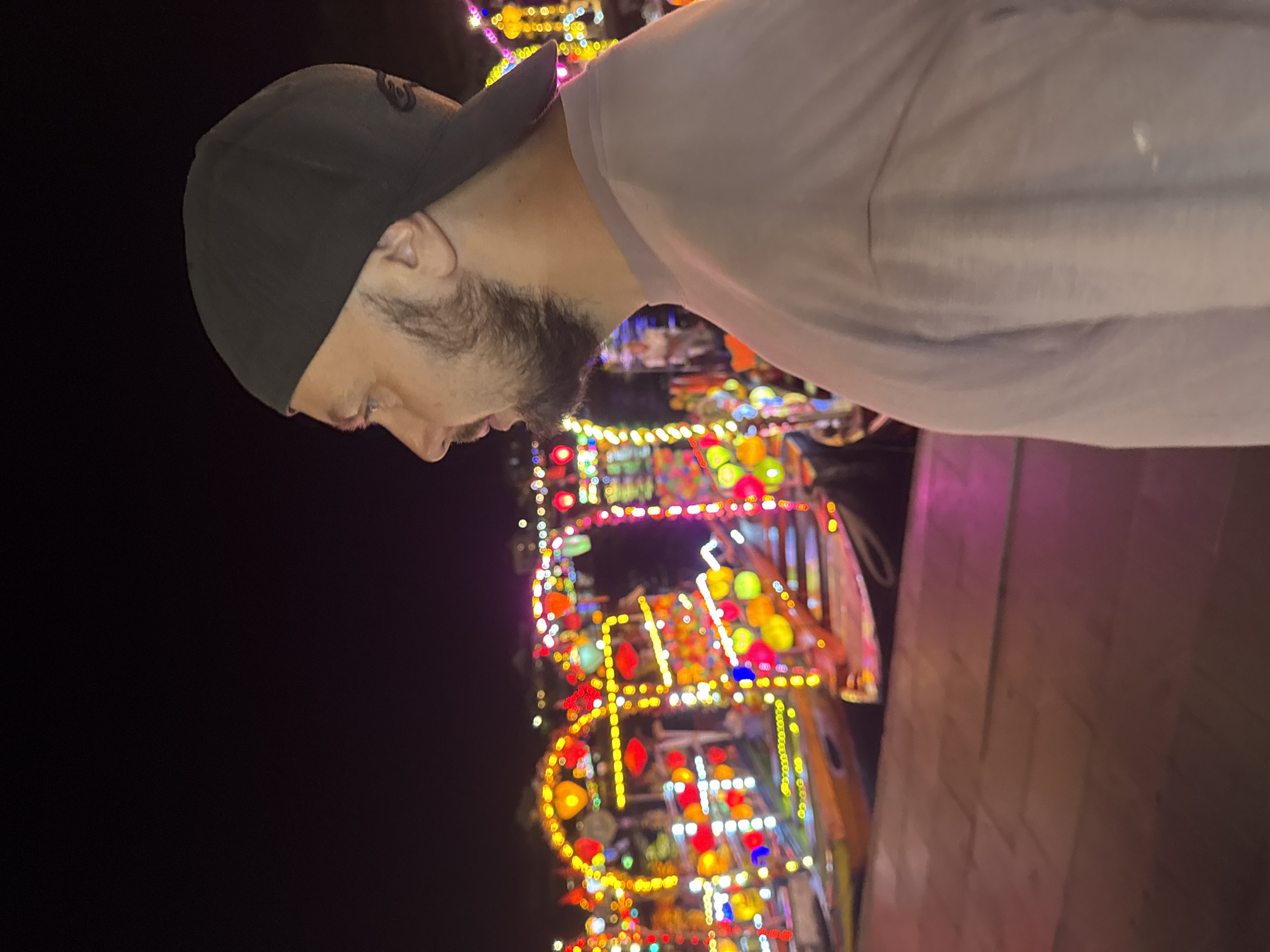 The image, taken in Hội An Ward, Vietnam, features a man wearing a black baseball cap and a light-colored shirt. He is oriented facing left with his head tilted downward. He has a beard. Behind him, a brightly lit carnival ride or structure is visible in the dark night. The ride is decorated with numerous colorful lights, primarily red, yellow, and blue. A brick-like structure is seen on the right side of the image.
