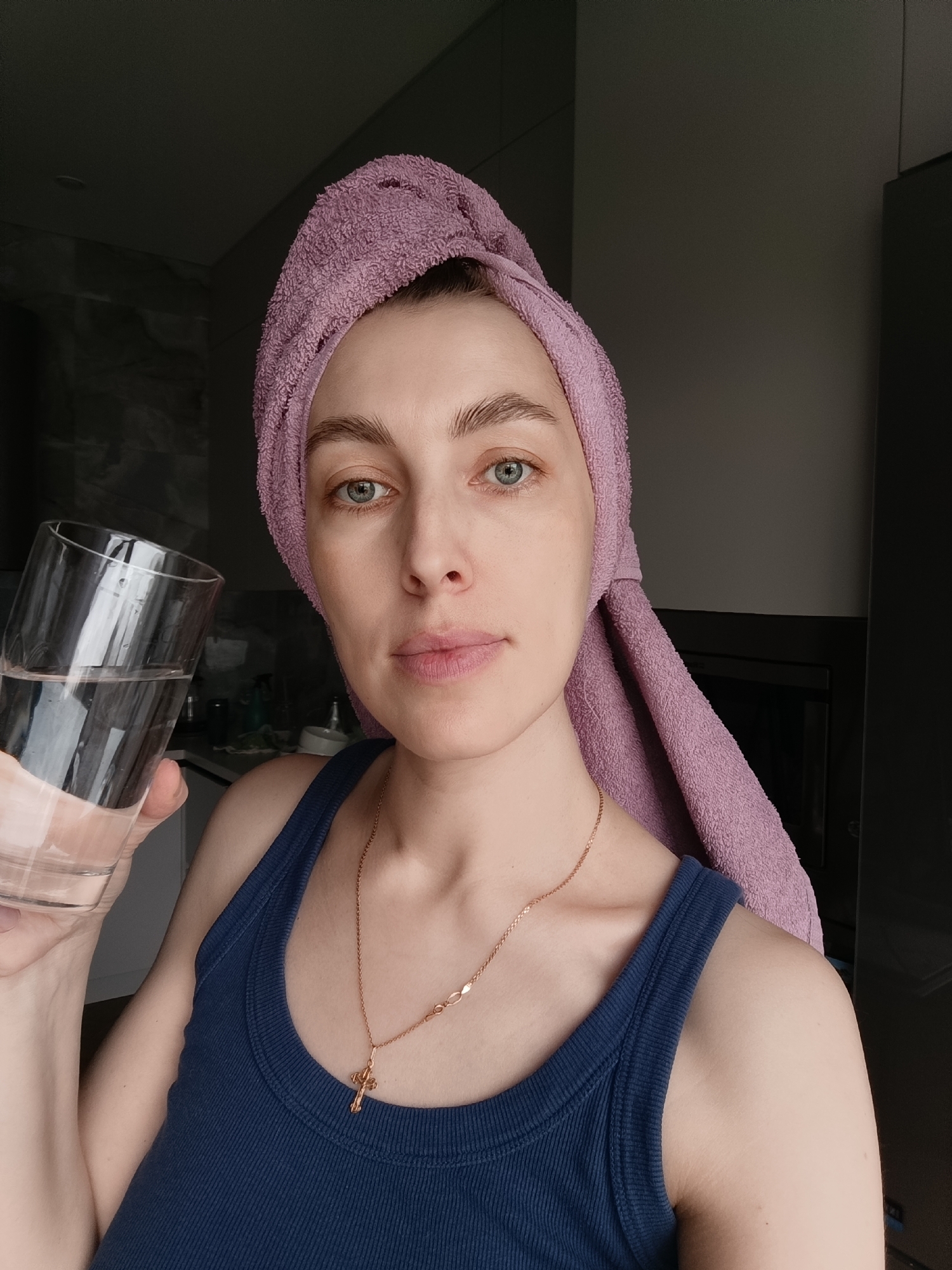 A fair-skinned woman with light blue eyes and a light pink towel wrapped around her head is holding a glass of water in her left hand. She is wearing a dark blue ribbed tank top and a gold necklace with a cross pendant. She appears to be indoors, possibly in a kitchen, based on the glimpse of cabinetry in the background. The scene is in the city of Chaikovsky, Russia.