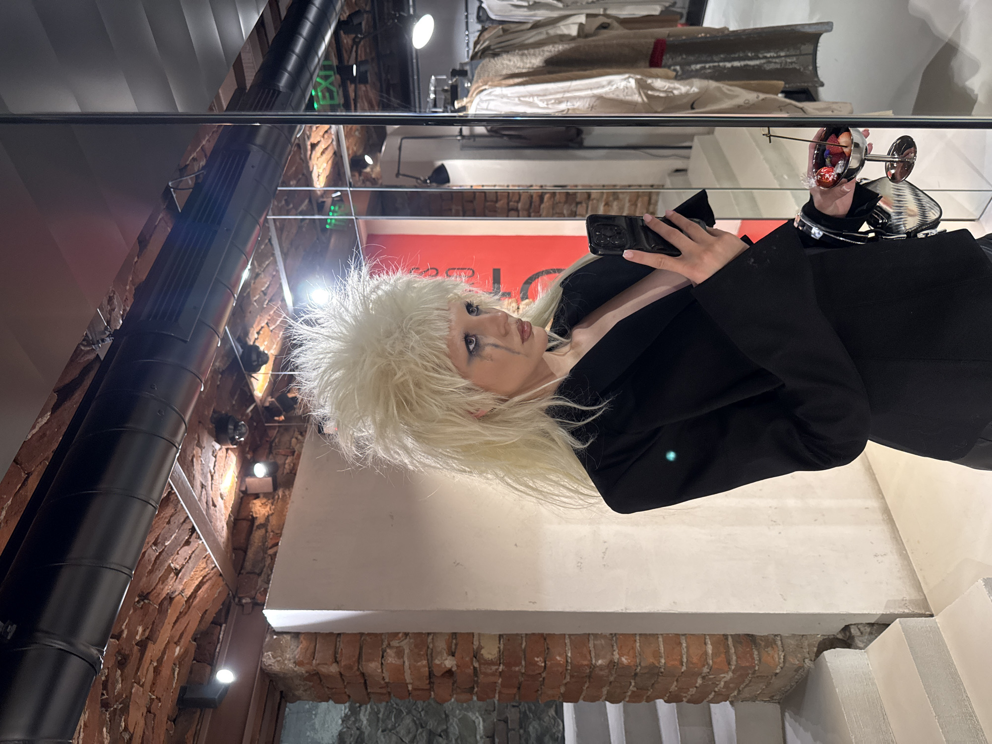 A female subject with stark, voluminous bleached blonde hair styled in a spiky, punk-rock aesthetic is depicted indoors in Sofia, Bulgaria, within a modern commercial establishment. Her face features dramatic black eye makeup streaked vertically down her cheeks. She wears a black blazer, open at the front, revealing her décolletage.
The subject is holding a black smartphone vertically in her left hand, angled as if taking a selfie or recording. In her right hand, she holds a small, silver-toned coupe glass containing dark red liquid, several berries (strawberries and blueberries), and a small wrapped candy, all pierced by a slender black stick. A small, metallic silver handbag hangs from her right wrist, alongside a metallic bracelet.
To the subject's immediate left, a pale, off-white wall adjoins an exposed red brick column, below which light-colored concrete stairs ascend. The setting appears to be a fashion boutique or gallery. The background to the right consists of a reflective surface, likely a mirror or glass wall. This surface reflects portions of the interior space, including an illuminated green "EXIT" sign, racks displaying various garments (some beige, some sequined), a dark modern floor lamp, and a red wall panel. The ceiling, visible both directly and in reflection, features exposed red brick arches, black industrial-style ventilation ducts, and recessed spotlights.