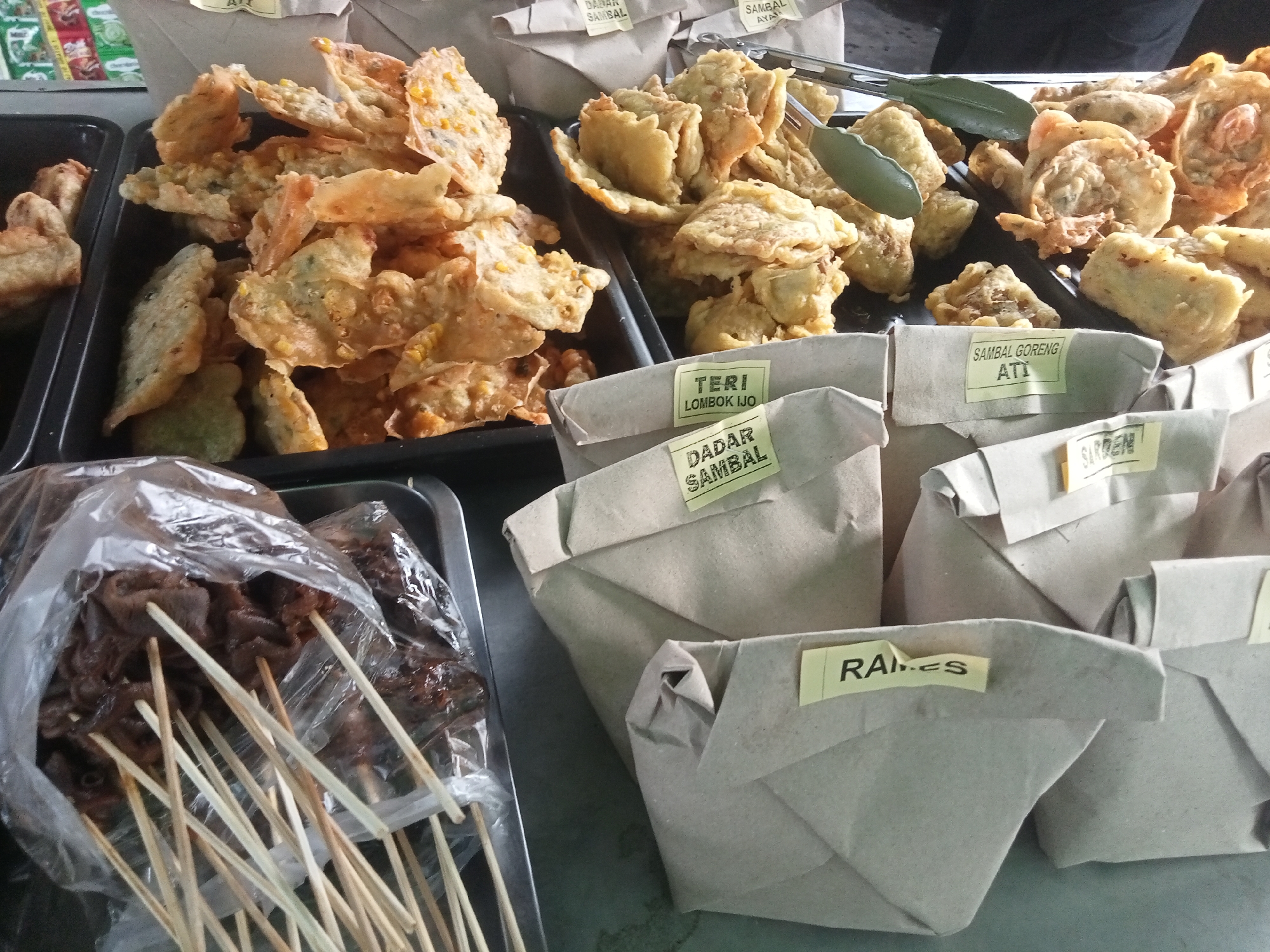 This is a close-up, high-angle shot of a food stall, likely in Semarang, Indonesia, displaying a variety of Indonesian snacks and dishes. In the foreground, a tray holds several skewers of dark, marinated meat wrapped in plastic. To the right, a row of paper-wrapped portions of food is arranged, each with a yellow label. The labels visible read "TERI LOMBOK IJO", "DADAR SAMBAL", "SAMBAL GORENG ATI", and "RA. ES". Behind these, a black tray overflows with various fried snacks, including what appears to be tempeh and corn fritters. In the background, partially obscured, are more paper-wrapped food items and glimpses of packaging with text. The overall impression is one of a bustling street food vendor ready for business.