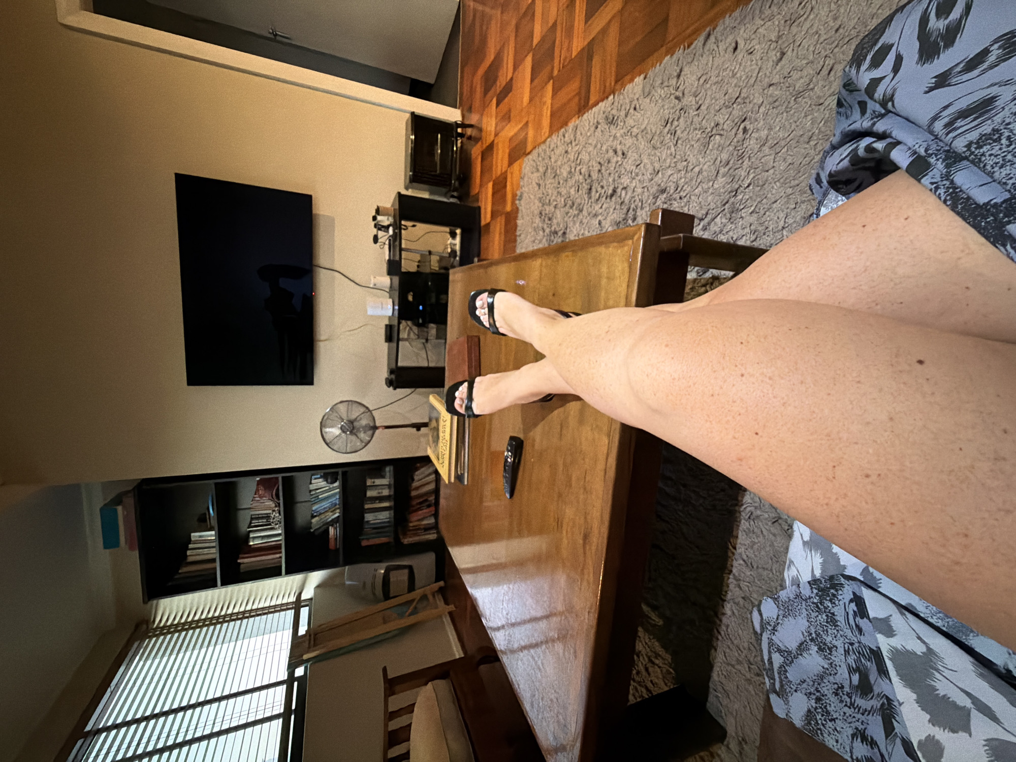 The image depicts an indoor scene, likely a living room. A person is seated, wearing a blue and white patterned garment, with their legs crossed over a wooden coffee table. The person is wearing black sandals. On the table, a TV remote and a book are visible. In the background, there's a television mounted on the wall, and a shelving unit containing books. A fan is positioned near the shelving unit. Sunlight streams in through a window with closed blinds. A grey rug is visible on the floor. The city of Cape Town, South Africa is not visibly discernible from the image.
