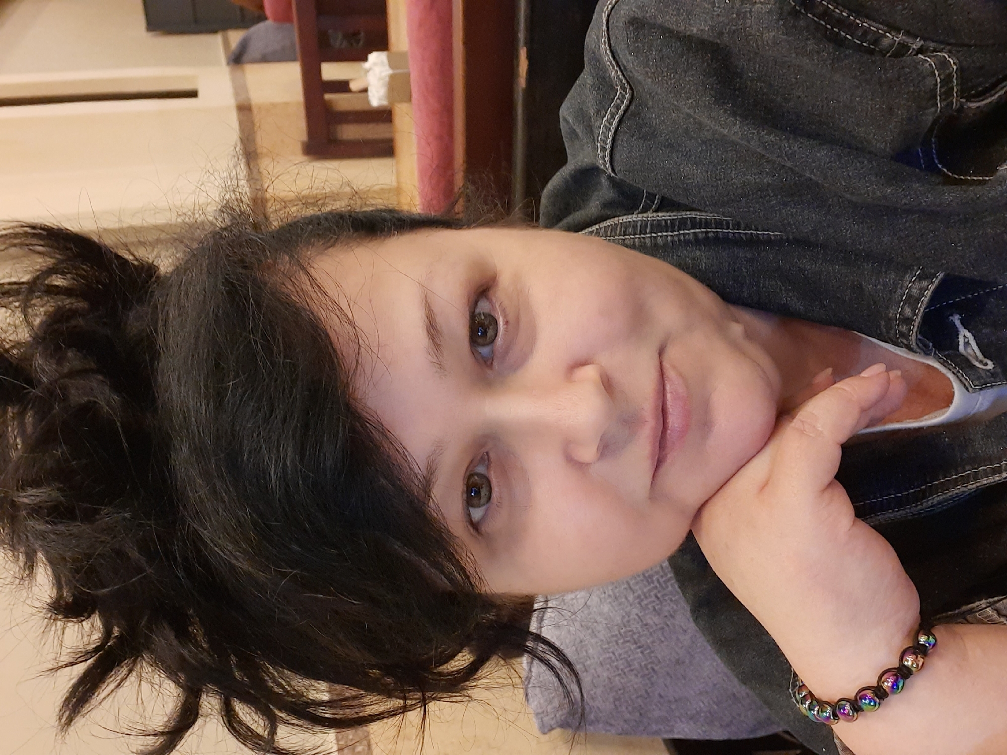 A close-up, slightly canted portrait depicts a woman with dark hair pulled up in a messy bun. Her skin is fair, and her eyes are hazel. She rests her cheek on her right hand, on which she wears a beaded bracelet with iridescent spheres. She is wearing a dark denim jacket over a white top. The background is blurred, but reveals glimpses of interior elements, including wooden furniture and a tiled floor. The location is unknown.