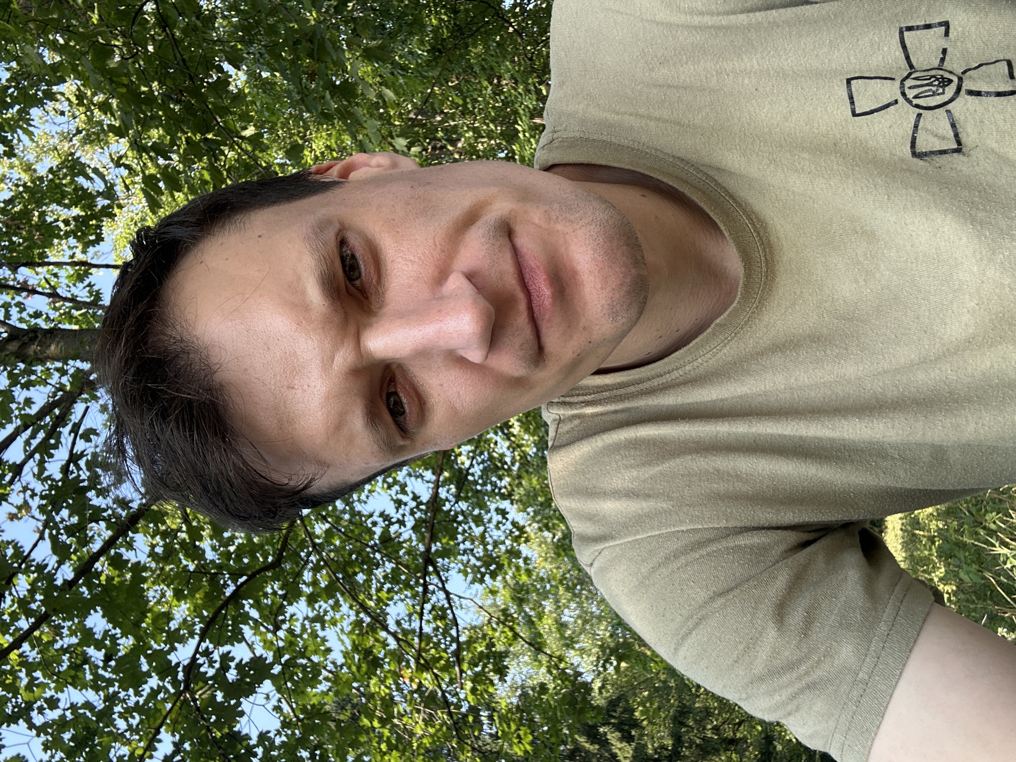 A man is pictured from the chest up, looking directly at the camera. He has short, dark hair and is wearing a light green t-shirt with a black symbol on the left breast. The man appears to be outdoors, with green foliage and a blue sky visible in the background. The location is identified as Makiv, Ukraine.