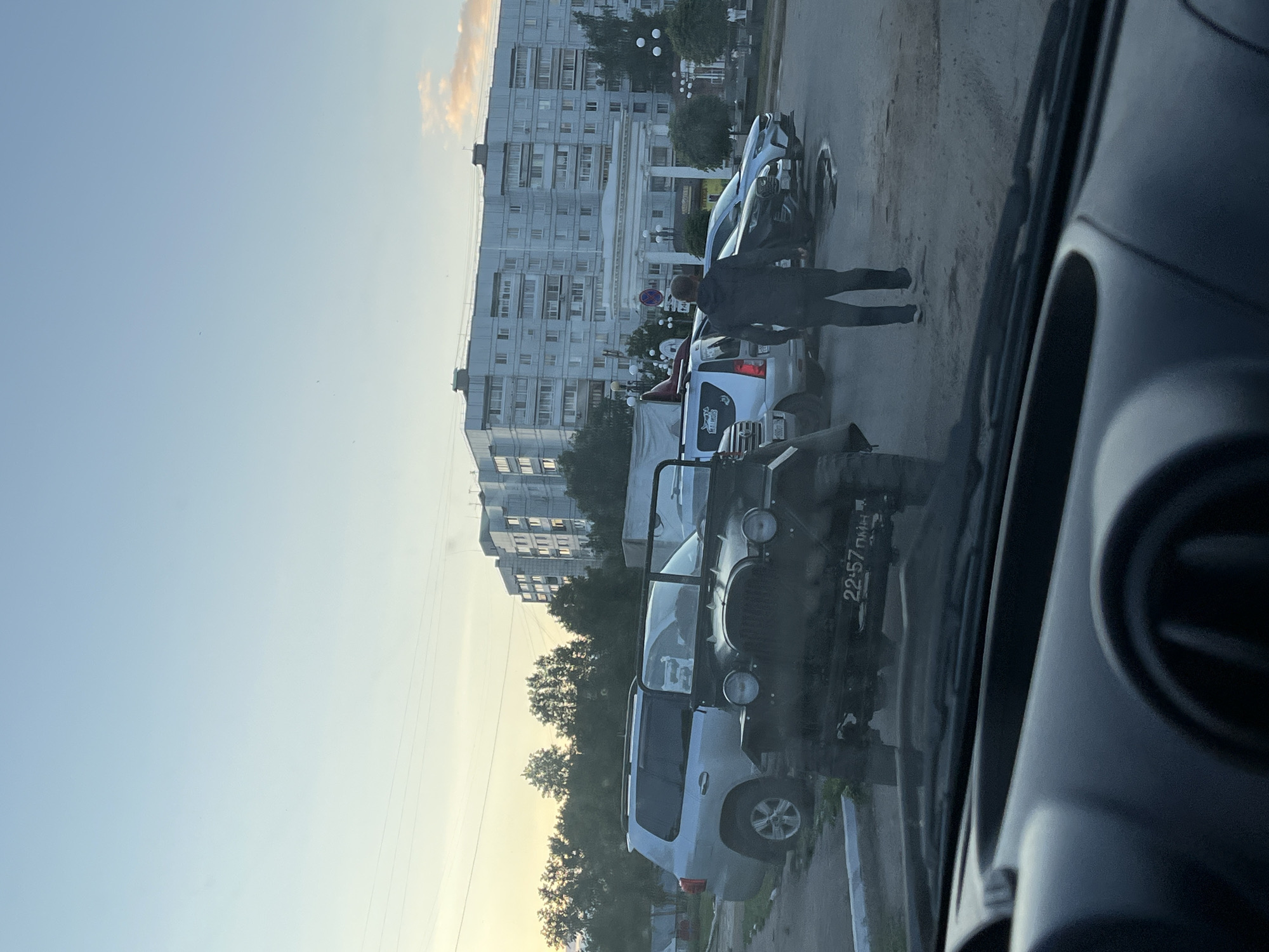 The image, taken from inside a vehicle looking out, captures an urban street scene in Chaikovsky, Russia, during what appears to be late afternoon or early evening. The right side of the frame shows the dark interior of the recording vehicle, including part of a dashboard and a circular vent.
In the midground, a dark green vintage-style off-road vehicle, possibly a UAZ, is prominently featured, parked near the curb. Its front passenger fender displays the text "22:57 пмн". Behind it, a silver SUV is parked, and further down the street, a white car and a dark sedan are visible. A motorcycle or scooter is also parked nearby. A person in dark attire (jacket and pants) stands on the street near the dark sedan and the white car, facing away from the camera. Another person is partially obscured near the white car. A blue circular road sign with a red border and a diagonal white line (indicating no stopping or parking) is visible behind the cars.
The background is dominated by large, multi-story residential apartment buildings typical of Soviet-era architecture, interspersed with green trees. The sky is a clear light blue, transitioning to a yellowish-orange hue near the horizon on the left, suggesting a sunset.