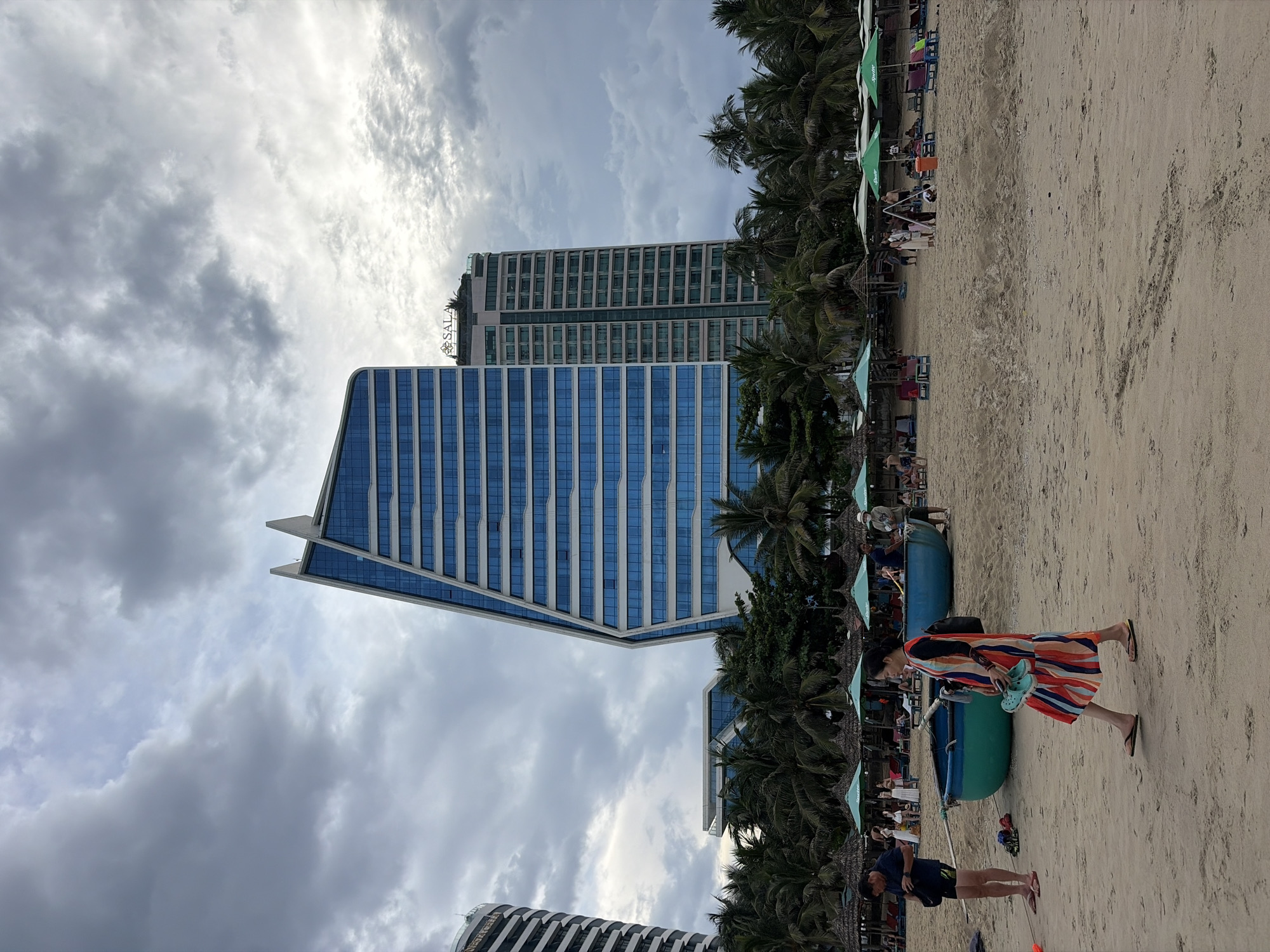 The image shows a beach scene in Da Nang, Vietnam, with a prominent, geometrically-shaped modern building featuring blue glass windows in the background. To the left, another tall building with a striped facade is partially visible. The beach itself is sandy, with several traditional Vietnamese fishing boats in teal and green colors resting on the sand. A woman in a colorful striped dress is walking across the foreground, holding a teal-colored shoe. In the background, there are rows of beach umbrellas and lounge chairs, and several palm trees line the edge of the beach. The sky is overcast with dramatic clouds. The overall scene depicts a resort area.