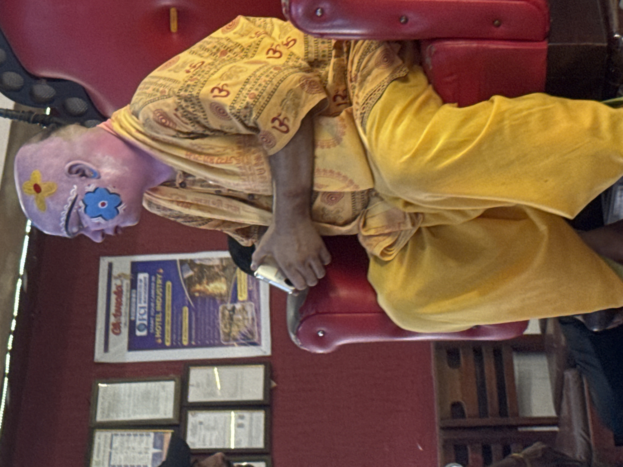A person with a shaved head, painted with a yellow flower and red center on the forehead and a blue flower design on the left cheek, sits in a large, red, high-backed chair. The person is wearing a yellow robe with an orange patterned trim and a yellow shawl draped over their shoulders. Their hands are clasped in their lap, holding a small silver object. The background is a red wall with framed posters on it. One poster is for "Oh India" and advertises the hotel industry. Wooden chairs are visible in the foreground to the left. The scene appears to be indoors, possibly a waiting area or a performance space, with the person dressed in what could be ceremonial or performance attire. The lighting is warm and slightly diffused.
