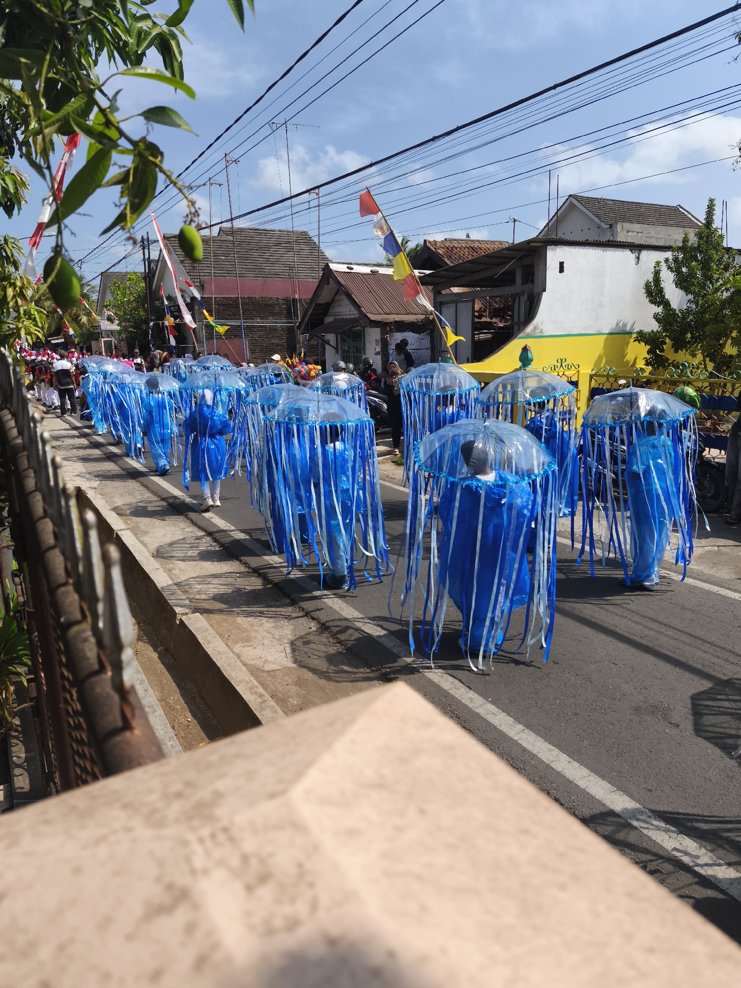 The image captures a street parade in Kutoarjo, Indonesia. A group of people, dressed in blue costumes resembling jellyfish with clear umbrella-like tops and dangling blue and white strips, are walking in formation on the right side of a paved road. The costumes cover them from head to toe, with the umbrellas obscuring their faces. Behind them, a crowd of spectators lines the sidewalk. In the background, buildings with varying roof styles and colors are visible, some adorned with Indonesian flags. Electrical wires crisscross the sky, and trees with green foliage can be seen. The scene suggests a festive occasion, likely a cultural event or celebration.