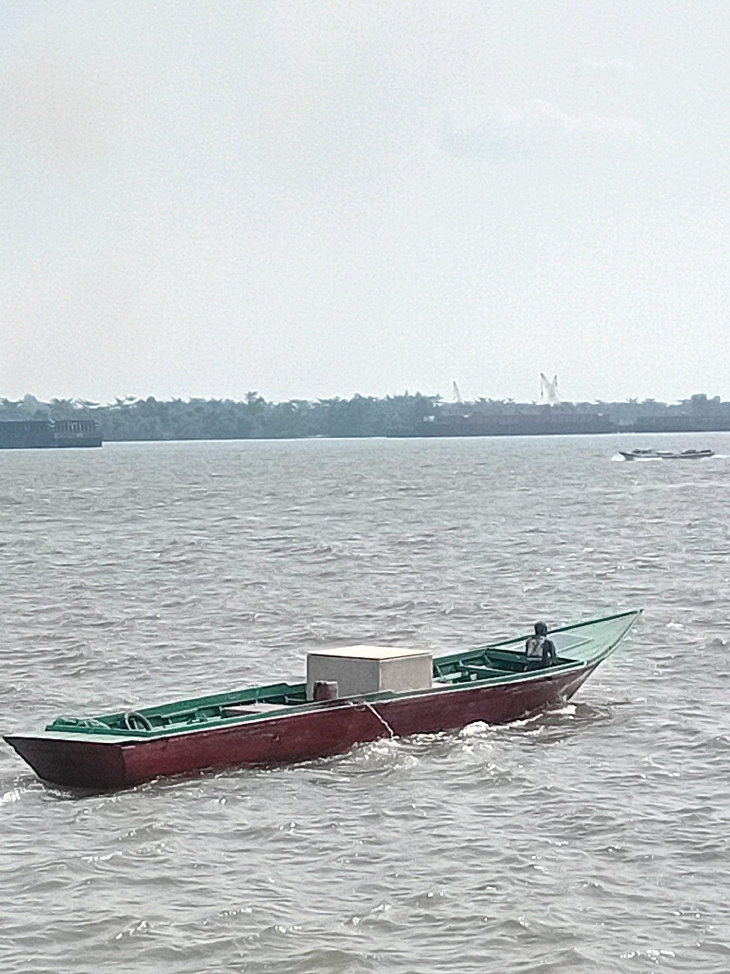 In Sungai Meriam, Indonesia, a narrow, red-hulled boat with a green interior is navigating a body of water. A lone individual is seated in the rear of the boat. A white rectangular object is positioned in the middle of the boat, secured by a rope. The water's surface is rippled, indicating movement. In the distance, a dense tree line forms the horizon, with structures and what appear to be cranes visible above the trees. Another smaller boat is moving in the distance to the right. The sky is overcast.