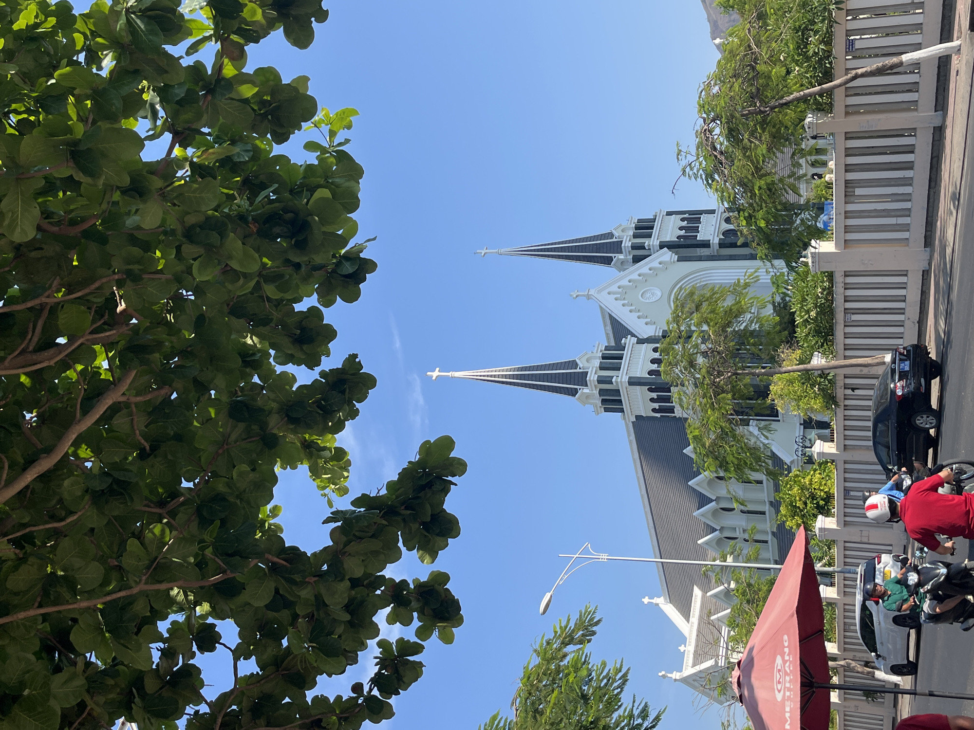 The image captures a view in the city of Bac Nha Trang Ward, Vietnam. A church with two prominent spires and white exterior details dominates the scene, viewed from a low angle partially obscured by a large tree with dense green foliage. The sky is clear and blue. A fence runs along the street bordering the church grounds. Several cars and motorbikes are present on the street, including one motorbike with a rider wearing a red shirt and white helmet. A red sun umbrella is partially visible in the lower right corner of the frame. A street light is placed near the church building.