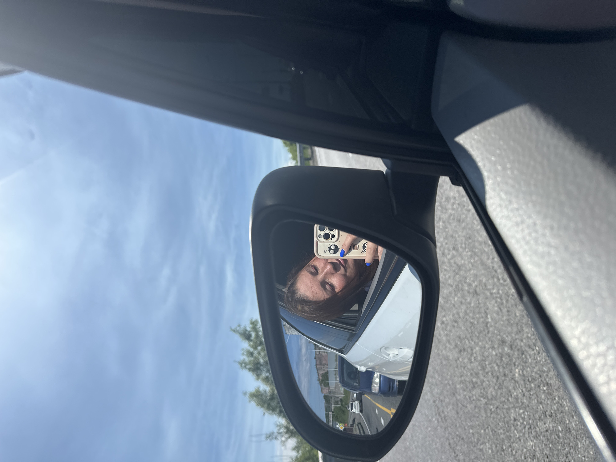 In the reflection of a car's side mirror, a young woman is visible. She is leaning out of the car window, holding a smartphone to take a selfie. Her eyes are closed, and her head is tilted upwards, suggesting a moment of enjoyment or relaxation. Her right hand, adorned with blue nail polish, is holding the phone. The phone case features a cartoonish design. The background reflected in the mirror shows a road with other vehicles, trees, and buildings. The overall scene appears to be a sunny day, with a clear sky visible. The image was taken from inside a vehicle, looking outwards, with the car's interior components, like the door frame and mirror housing, visible in the foreground. The location is identified as the city of Krutoberegovoy, Russia.