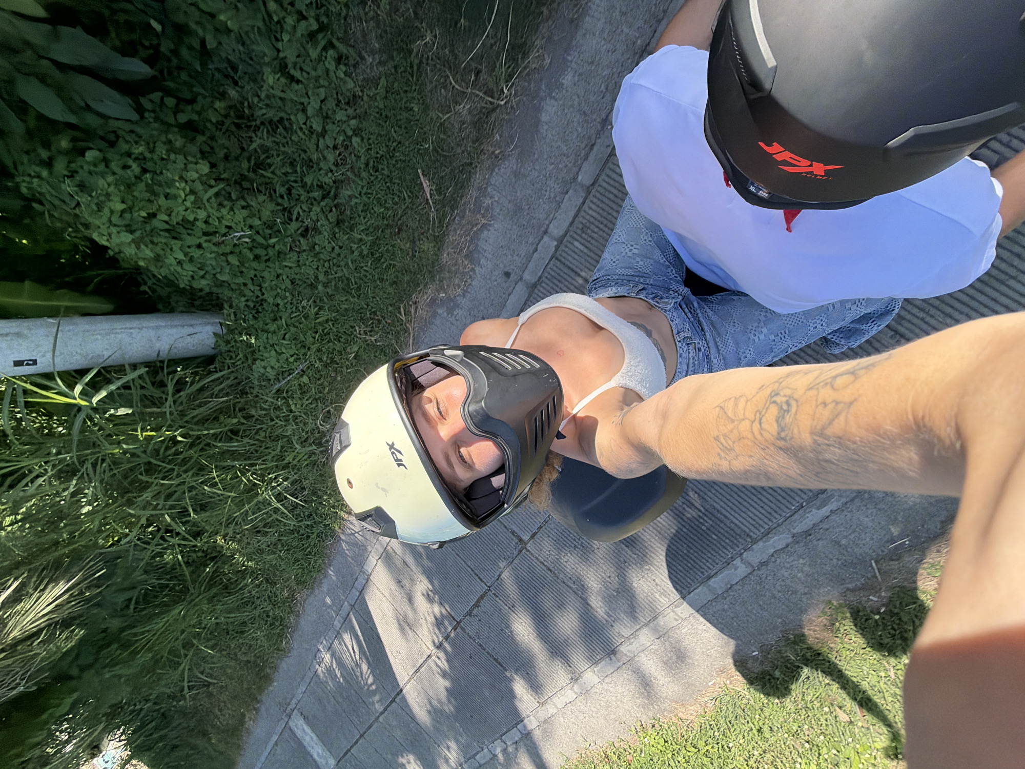 A top-down, eye-level photograph depicts two people wearing helmets, likely riding a moped on a street in North Kuta, Indonesia. The person on the left is wearing a cream-colored and black helmet with the letters "JPX" visible. The person on the right has a similar helmet, predominantly black with the "JPX" logo in red. The person on the left is wearing a white top and light blue jeans with patterns. The person on the right is wearing a white shirt. The photographer's arm, with tattoos, is visible. The ground appears to be paved with interspersed sections of green grass and plants. A metal pole and additional vegetation are seen in the left background. The scene is bathed in sunlight, casting shadows.