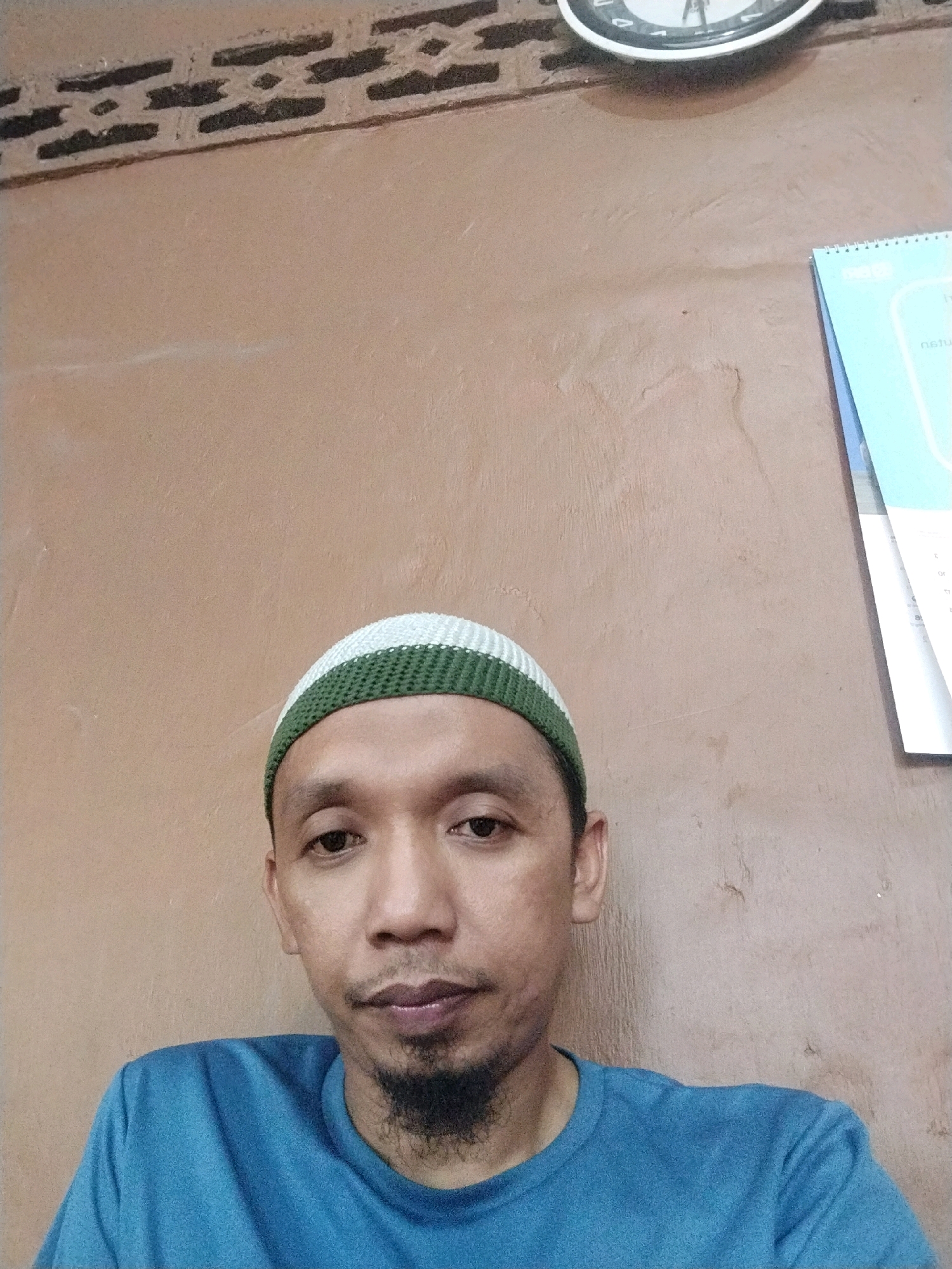 A man is wearing a blue t-shirt and a white and green kufi. He has a beard and is looking at the camera. In the background, there is a tan-colored wall, a clock on the wall, and a light blue calendar hanging on the wall. The scene appears to be indoors in a residential setting. This photograph was taken in Tanjung Marwo, Indonesia.