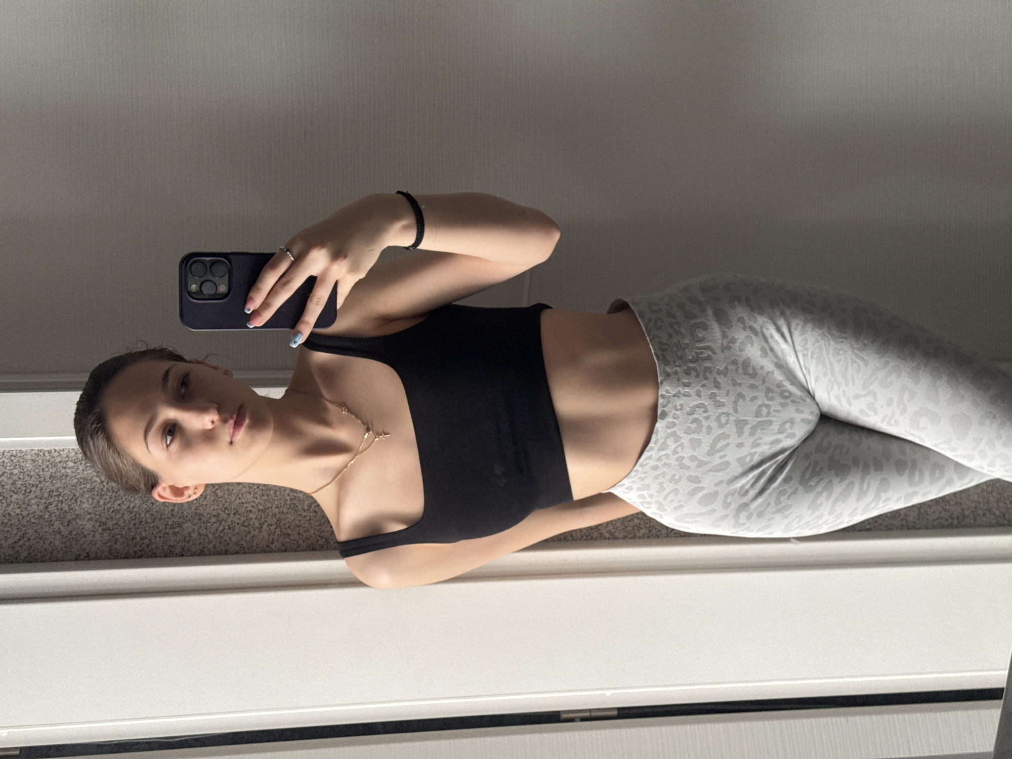 The image shows a young woman indoors taking a selfie. She is wearing a black sports bra and grey patterned leggings. She is holding a smartphone in her left hand, which is likely being used to take the picture. She has fair skin, brown hair pulled back, and is wearing jewelry, including a necklace and ring.
There's no information in the image that indicates the location of Sofia, Bulgaria. The environment appears to be inside a room with carpeted flooring and a white wall or a doorframe with a painted wall behind it.