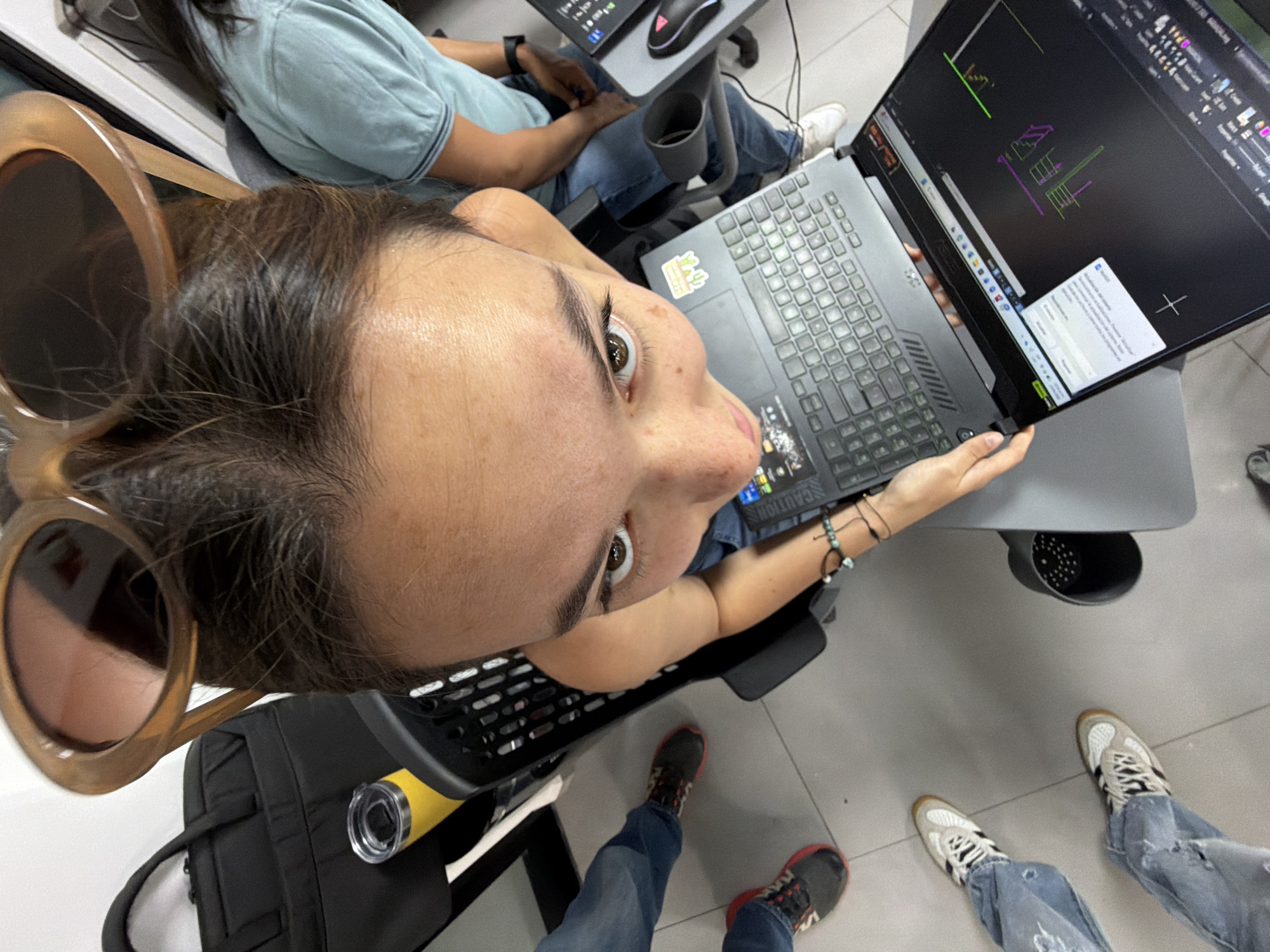 An eye-level, aerial shot depicts a woman holding a laptop in a room. She has light skin, brown hair, and wears large, round sunglasses. Her torso is visible, clad in a blue shirt. She's seated in a black chair with a black backpack at her side, a yellow water bottle placed inside the backpack's pocket. The laptop screen displays a CAD program with technical drawings. In the background, another person is seated with a laptop and mouse. Two pairs of feet, wearing different shoes, are visible near the bottom of the frame, suggesting additional people in the space. The floor is a light gray tile. The location is likely an office or educational setting in San Andrés Cholula, Mexico, though this cannot be definitively determined from the image.