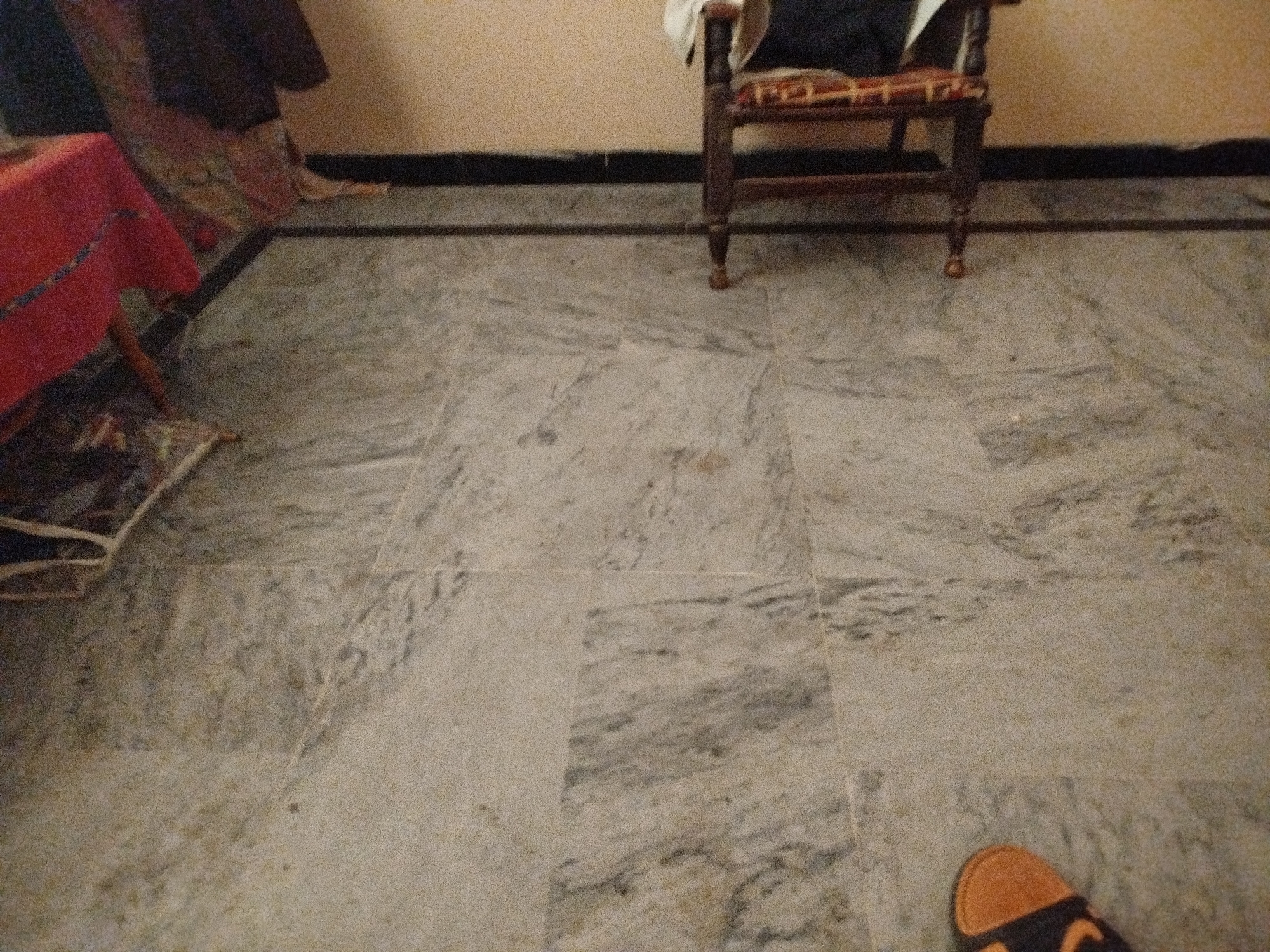 The image depicts an indoor scene with a marbled floor, likely in Lassan, Pakistan. In the background, a portion of a wooden chair with a woven seat and a white cloth draped over it is visible. To the left, a table covered with a red tablecloth and a patterned rug on the floor are partially shown. A dark band runs along the base of the wall, separating it from the floor. In the foreground, the tip of an orange and black sandal is visible on the floor. The lighting suggests an indoor environment, possibly a residential or waiting area.