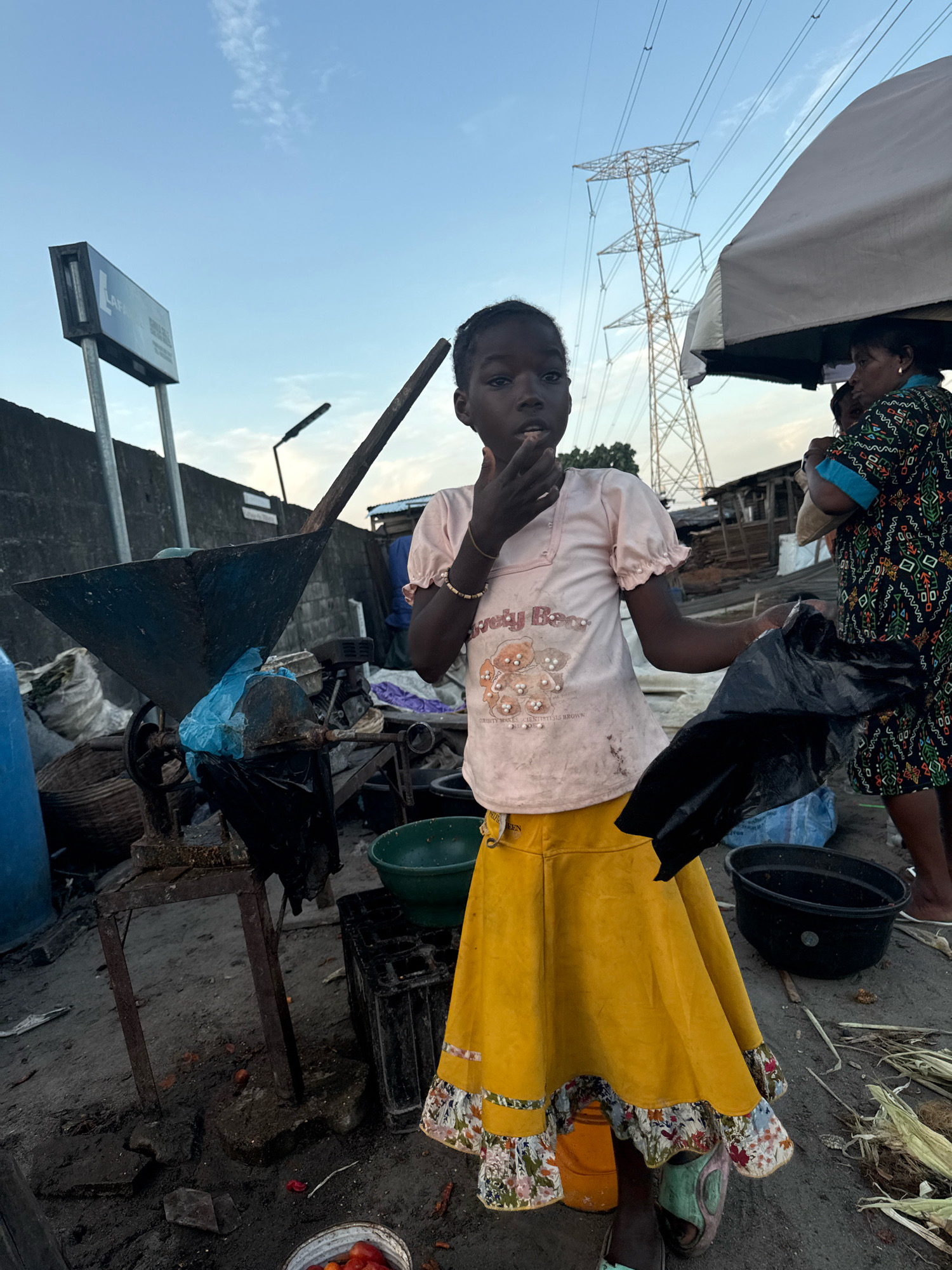 In Ado, Nigeria, a young girl stands in an outdoor market, wearing a light pink t-shirt with a graphic of cartoon bears and a bright yellow skirt with a floral hem. She has her right hand to her mouth, as if eating or tasting something, and holds a black plastic bag in her left hand. Behind her, a woman in a patterned dress is partially visible, also holding a bag. The background reveals a utilitarian scene with a large industrial-looking machine, various containers, and sacks of materials. Towering high-voltage power lines dominate the sky above, indicating the presence of significant electrical infrastructure. The ground is uneven and appears to be dirt or compacted earth, littered with some debris. The overall impression is a busy, functional marketplace environment with clear signs of everyday commerce and life.