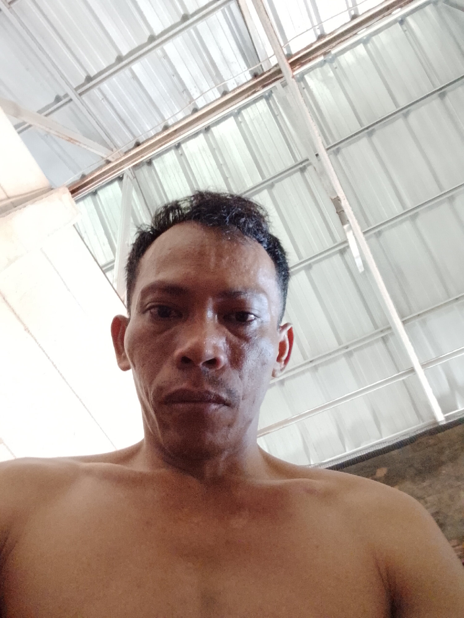 The image is a close-up selfie of a shirtless man with short, dark hair. The background shows a metal roof structure. The man appears to be middle-aged with weathered skin. The location is possibly in Bukit Ketok, Indonesia.
