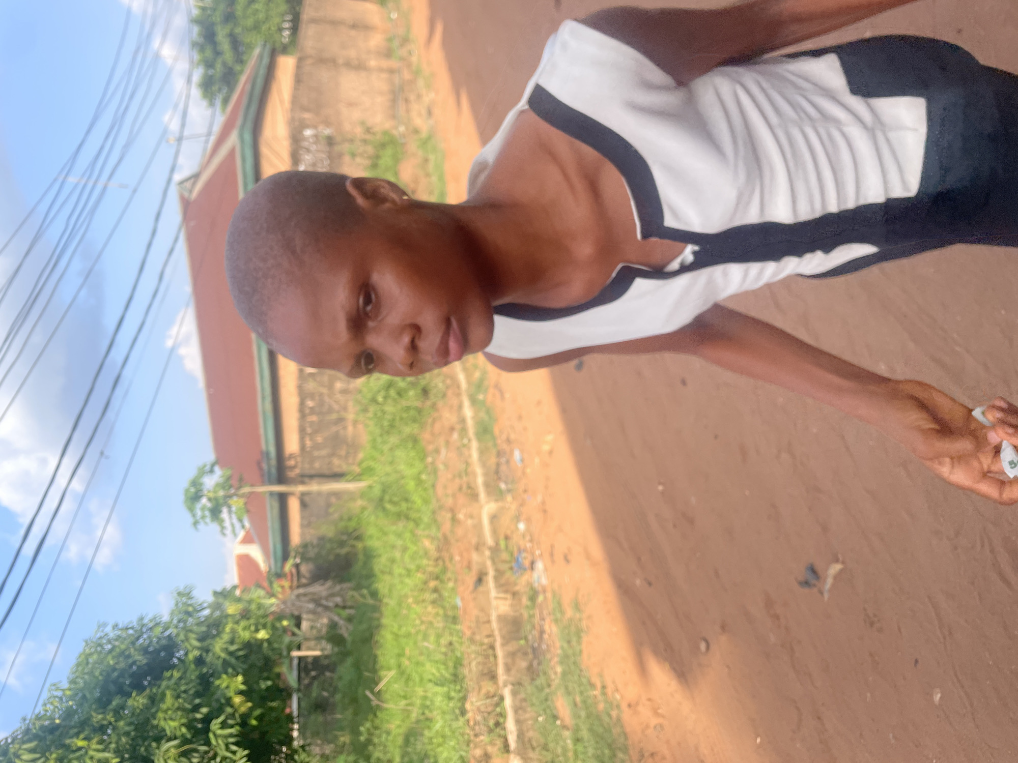 A close-up shot shows a Black person with closely cropped hair, possibly a buzz cut, standing on a dirt road or path in Nkpor, Nigeria. They are wearing a white sleeveless top with a dark trim around the neck and armholes, and dark-colored bottoms. The person is looking towards the camera. The background includes green foliage, a building with a light-colored wall and dark roof, and overhead electrical wires. The ground is reddish-brown. The photograph appears to have been taken in daylight. A wrapper/package is visible in the right hand.