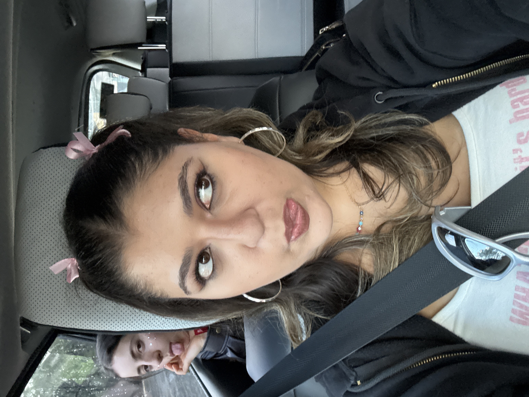 A woman is sitting in a car wearing a black jacket over a white top. She has light brown hair parted in the middle, hoop earrings, and a seatbelt across her chest. In the reflection of the window, a person is sticking their tongue out. The location appears to be Mexico City, Mexico.